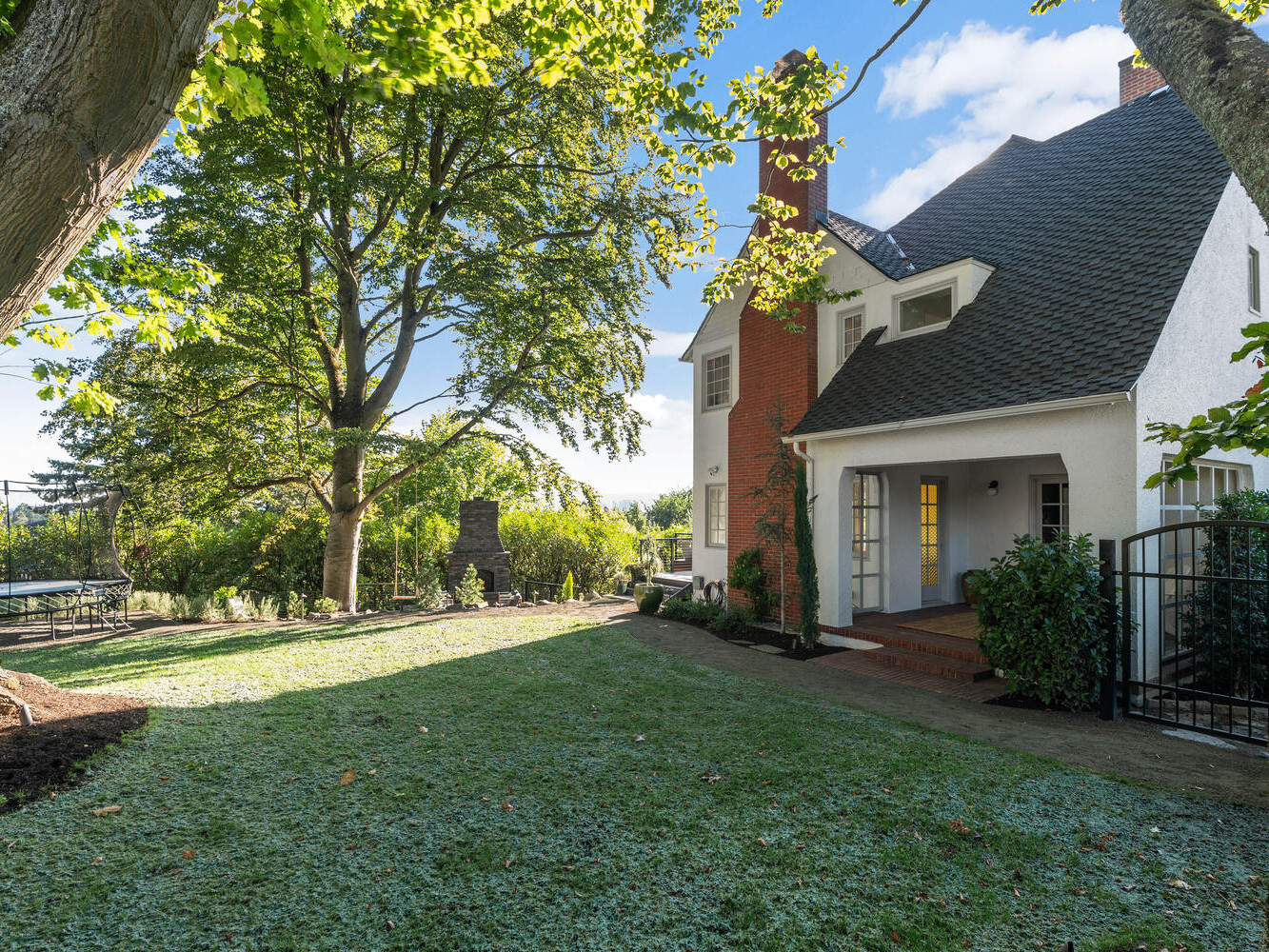 A charming house with a sloped roof nestled in a lush Portland, Oregon garden. Large trees provide shade over the lawn, and a stone pathway leads to the entrance. A tall brick chimney accents the white exterior of this delightful home.