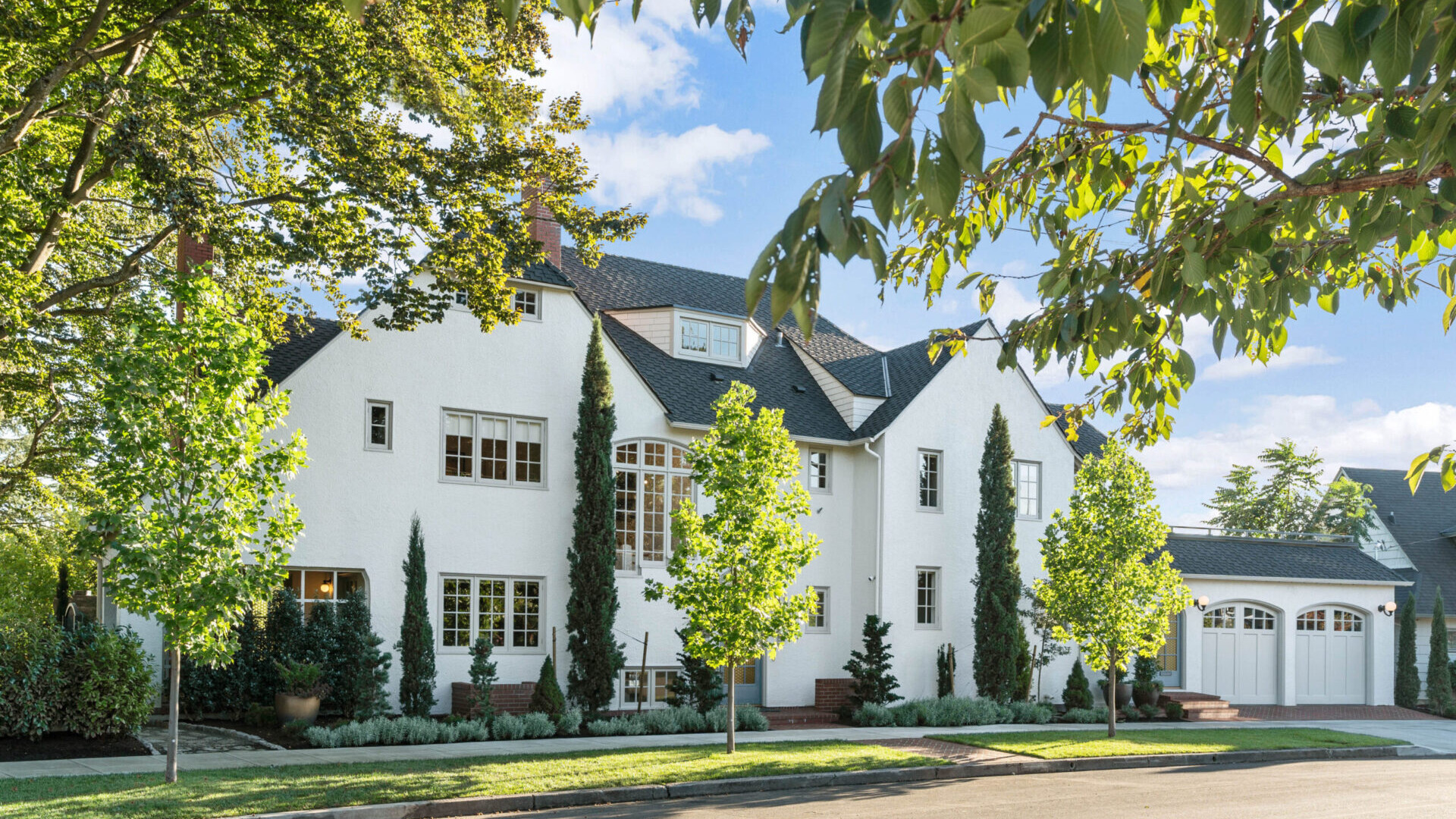 A large white house with multiple windows, surrounded by green trees and a manicured lawn. The clear blue sky enhances the serene suburban setting, showcasing Portland Oregon real estate at its finest. A driveway leads to a white double garage on the right side.