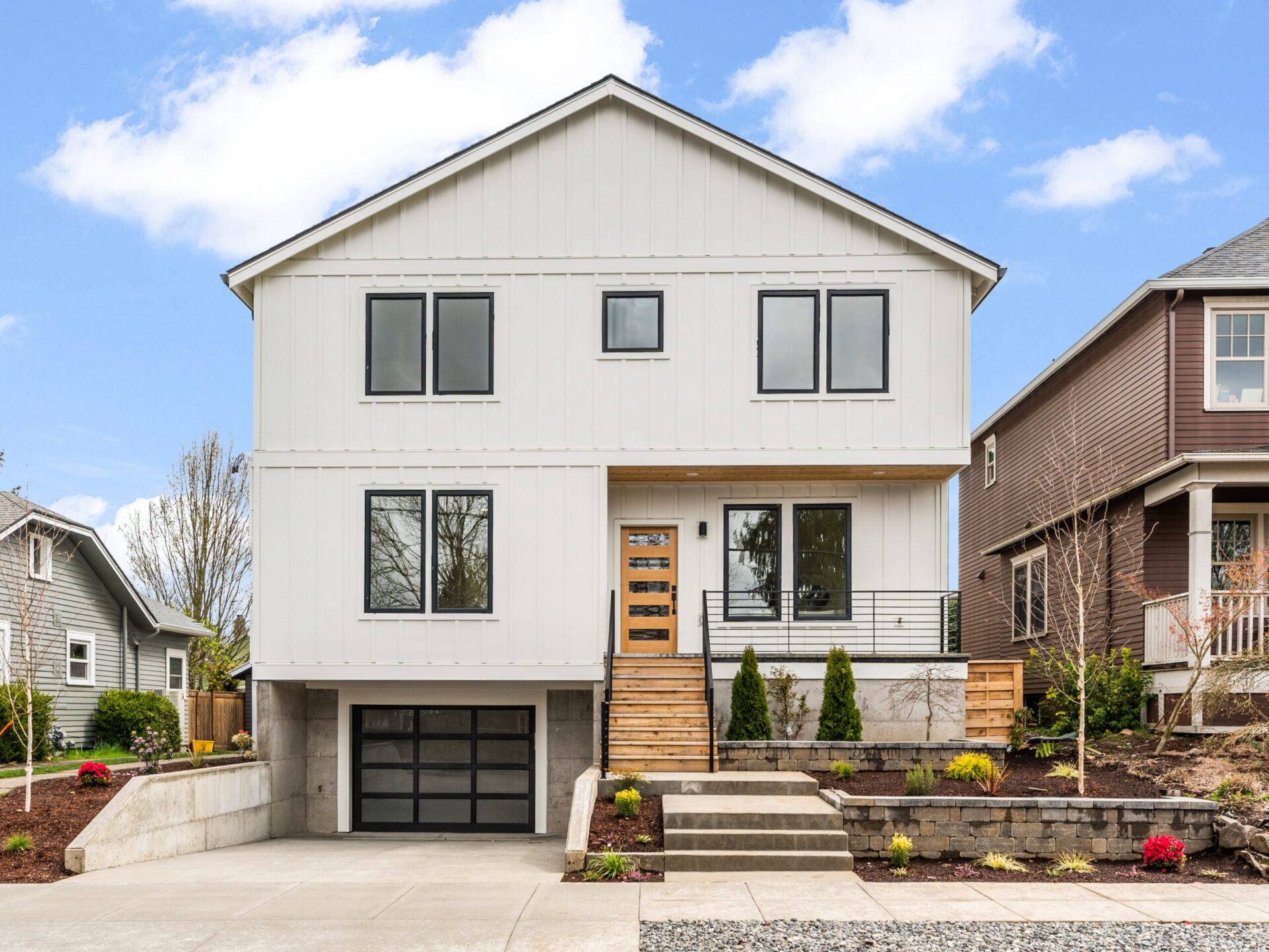 A modern, two-story white house in Portland with a black garage door, large windows, and a wooden front porch. The front yard features neatly landscaped plants and a concrete driveway. Discover it under the clear blue sky with guidance from your top Portland Realtor.
