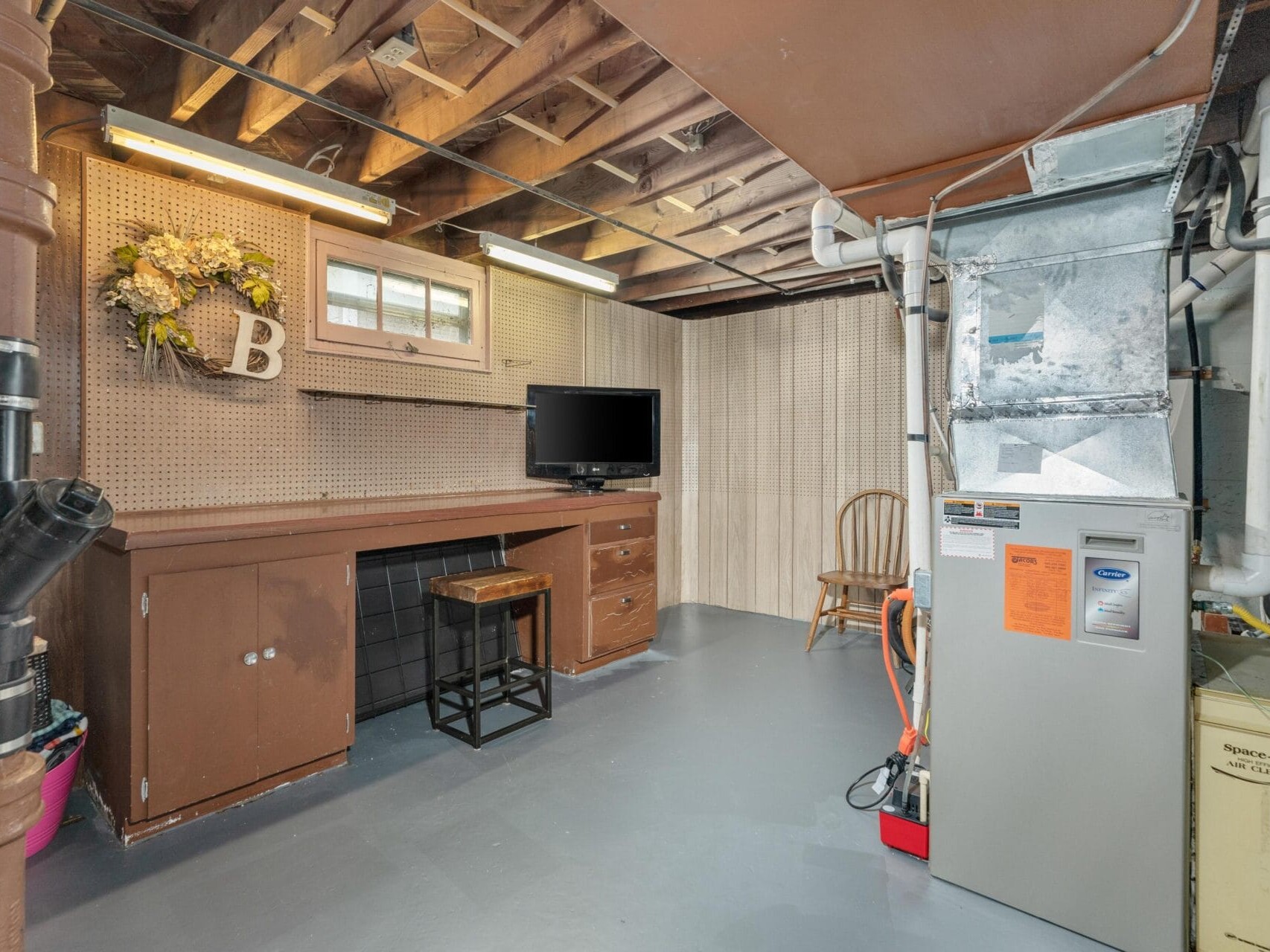 A basement with exposed wooden beams, featuring a workbench with a small TV, chair, and ventilation system. A decorative wreath with the letter B hangs on the wall, and the floor is painted gray. The space is well-lit.