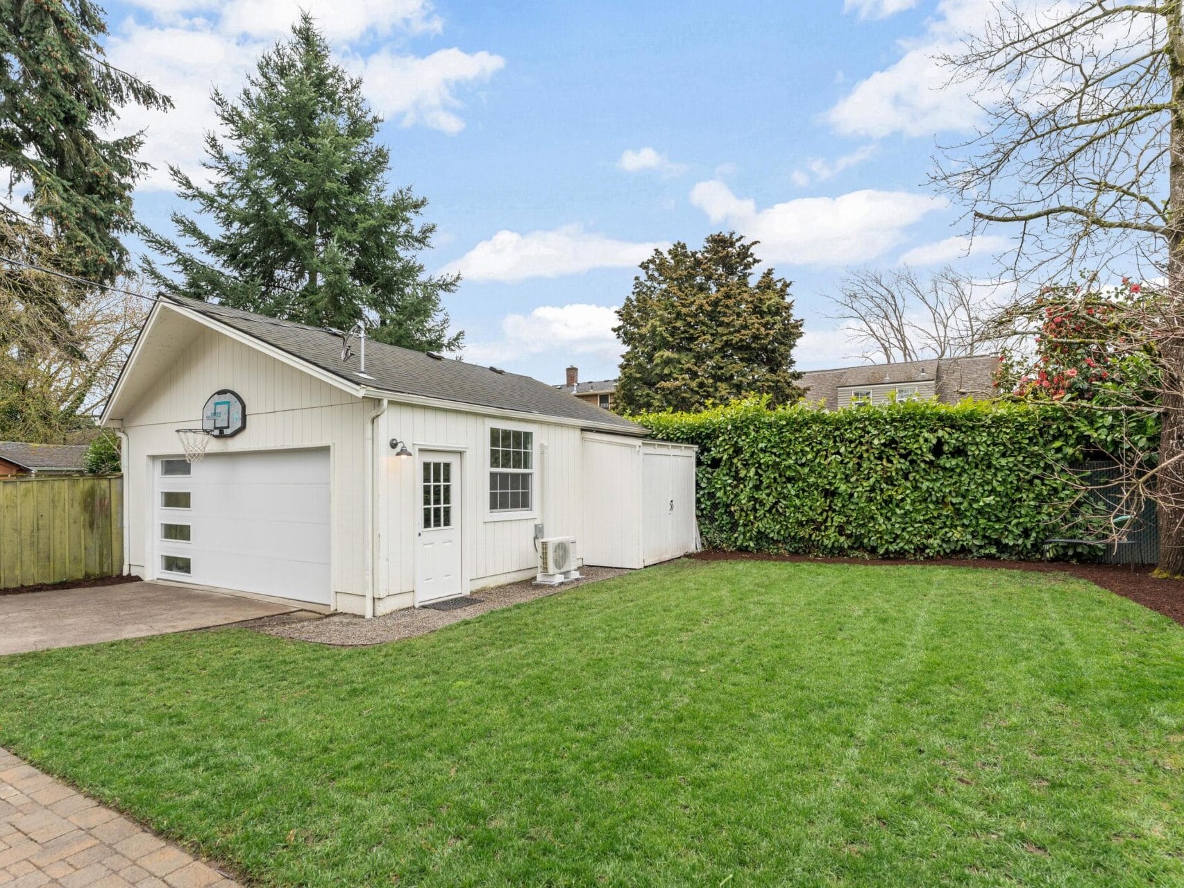 A white garage with a basketball hoop is surrounded by a well-maintained lawn and trees. A wooden fence lines the property, and a clear blue sky is visible above.
