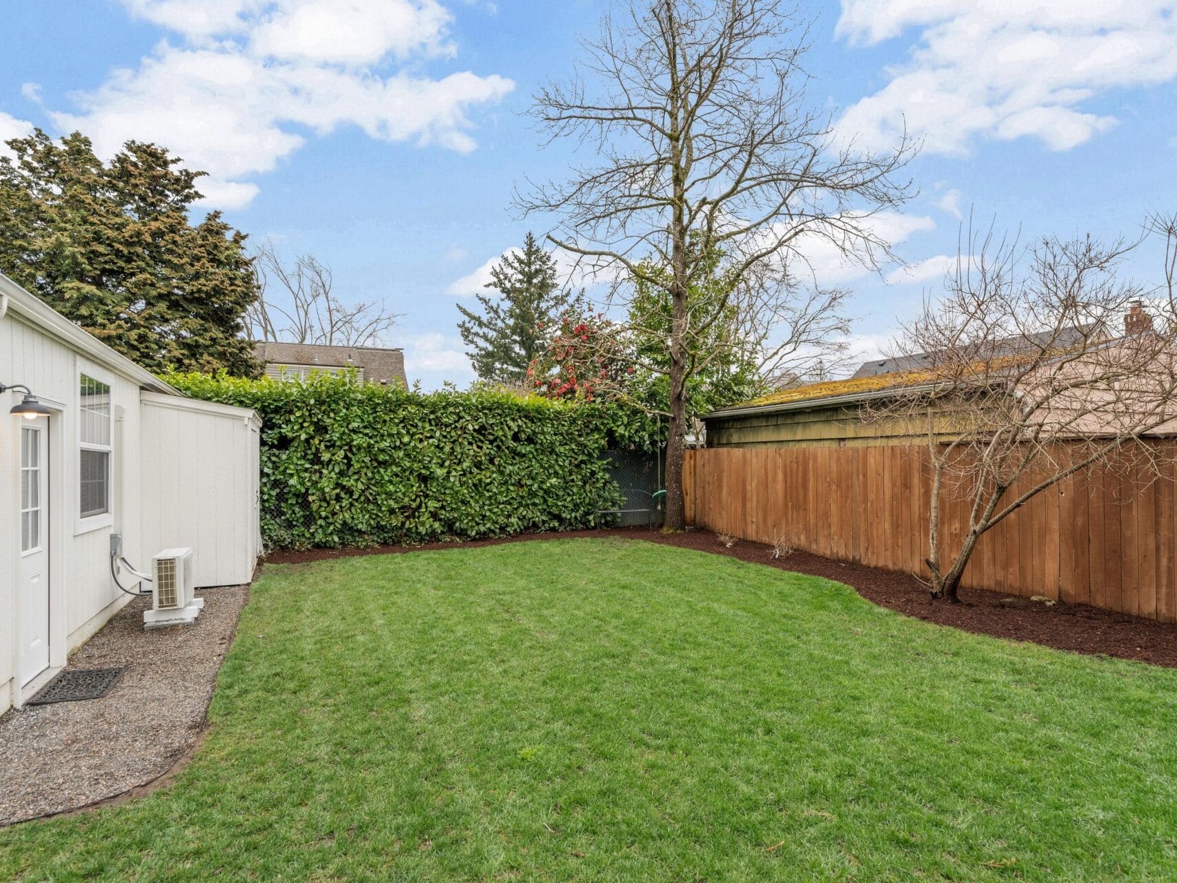 A backyard with a green lawn bordered by a wooden fence on the right and a hedge on the left. A small building with a door and windows is on the left side of the image. The sky is partly cloudy.