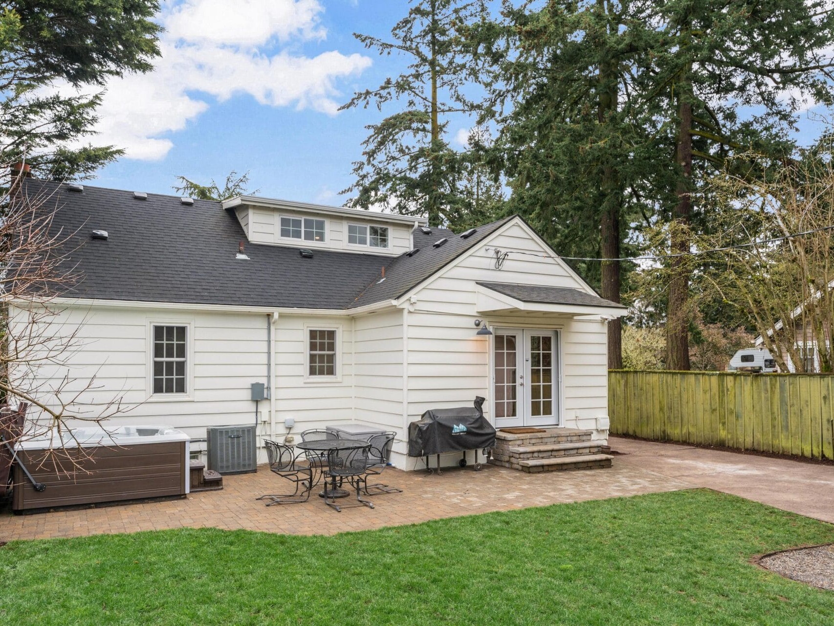 A white house with a pitched roof and dormer windows. The backyard features a stone patio with a dining set and a grill. A hot tub is on the left, surrounded by trees and a wooden fence under a partly cloudy sky.