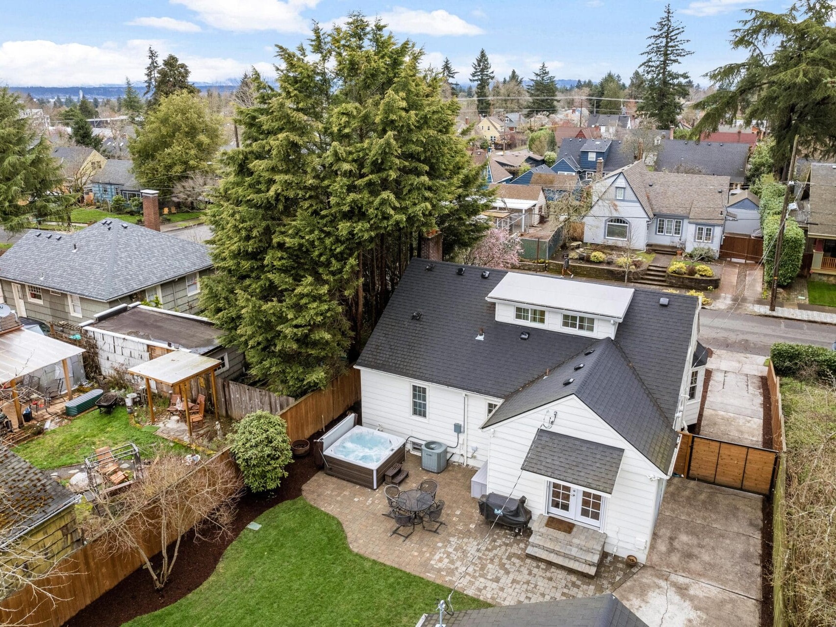 Aerial view of a suburban neighborhood featuring a white house with a black roof. The backyard has a patio with seating, a hot tub, and is surrounded by trees and a fence. Other houses and trees are visible in the background under a partly cloudy sky.