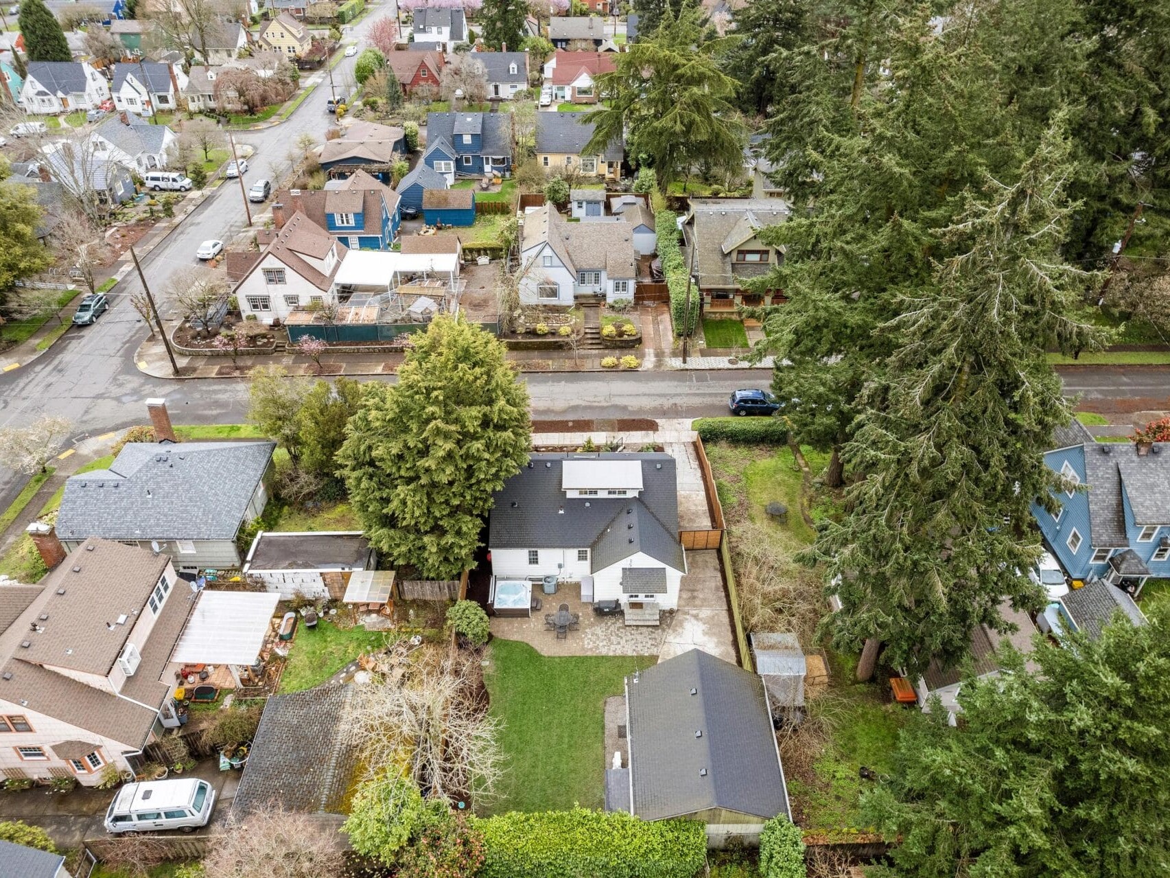 Aerial view of a suburban neighborhood with various houses surrounded by trees. The scene includes roads, yards, and a mix of evergreen and deciduous trees, creating a lush environment. The homes are arranged closely, with some having backyard spaces.