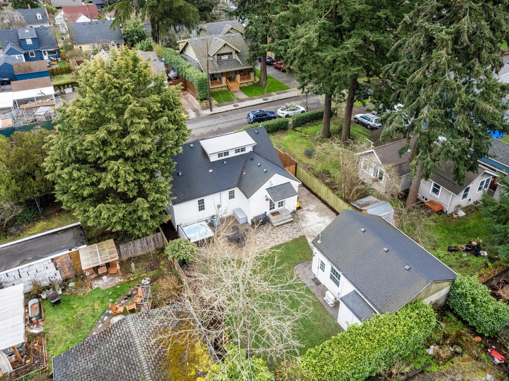 Aerial view of a residential neighborhood featuring several houses surrounded by tall trees. A white house is central in the image, with a backyard and patio. Streets and parked cars are visible, and greenery is abundant throughout the area.