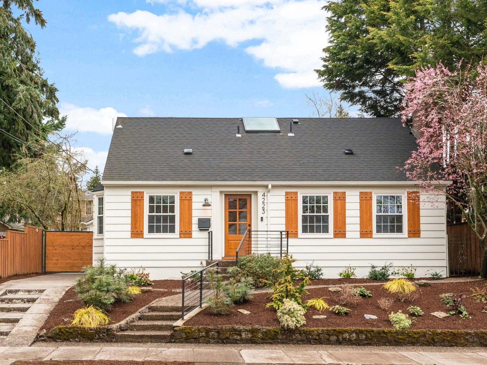 A charming white house with a black roof and orange shutters, surrounded by small shrubs and blooming trees. A paved walkway and a few steps lead to the front door. The sky is partly cloudy.