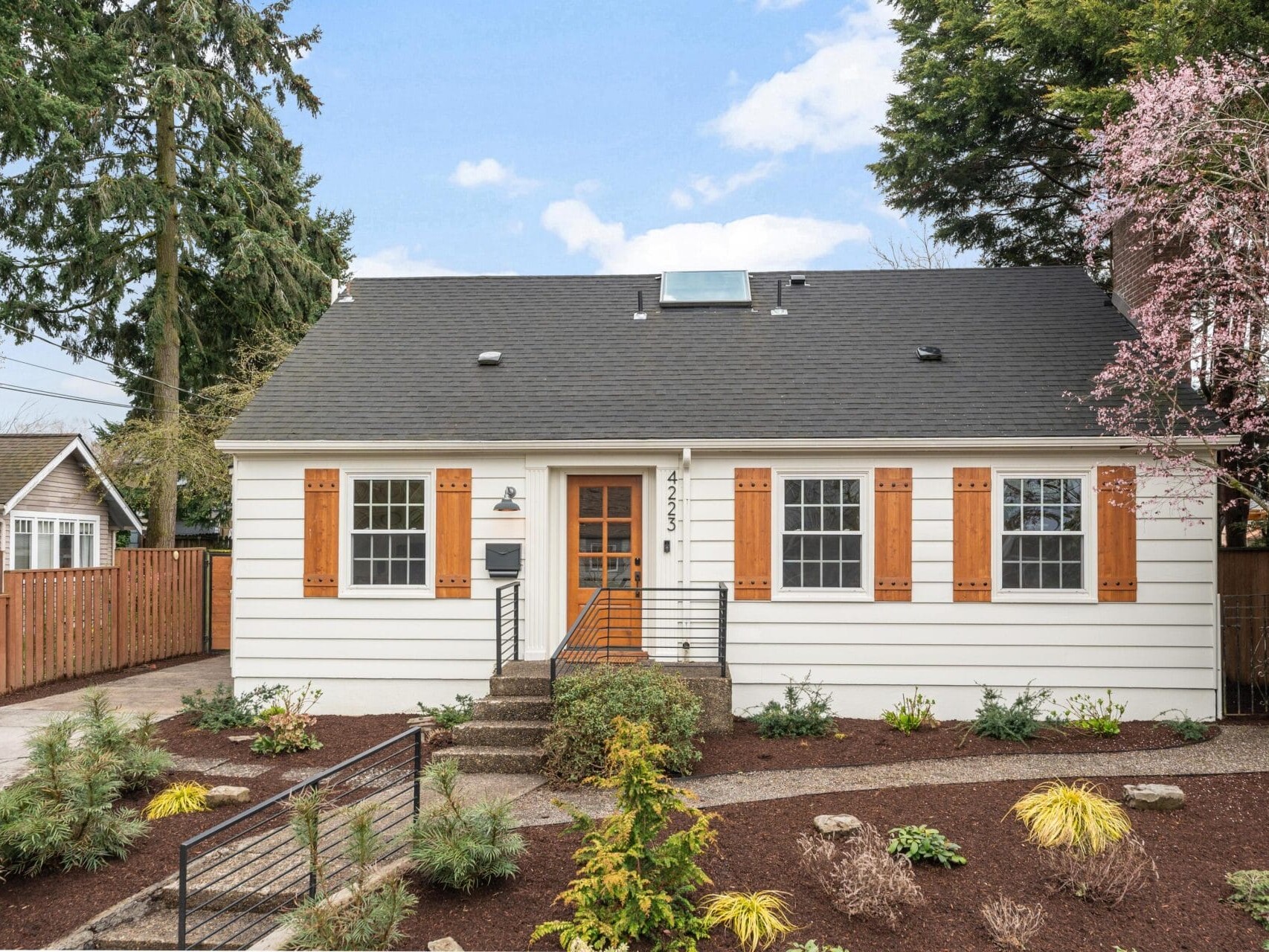 A charming white house with a dark roof, orange shutters, and a front garden with shrubs and mulch. A small path leads to the orange front door. Trees and a wooden fence are in the background.