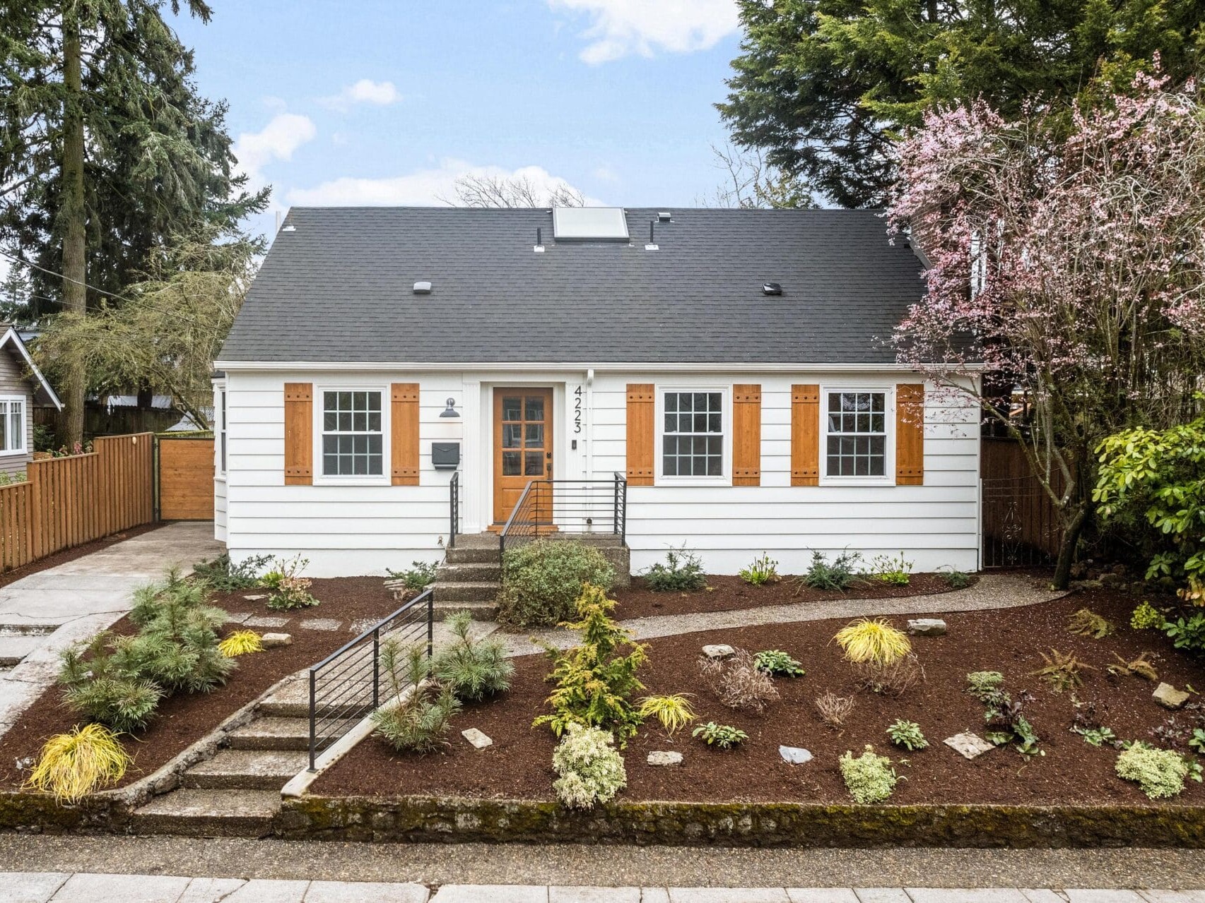 A white house with wooden shutters sits behind a freshly landscaped front yard featuring various plants and shrubs. Theres a small stairway leading up to the door, and large trees frame the house.