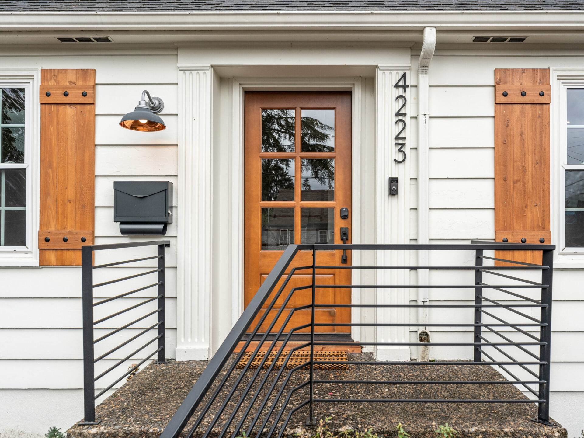 A modern front entrance of a house in Portland, Oregon real estate, featuring a wooden door with glass panels, flanked by wooden shutters on two windows. A black mailbox is mounted on the left, and the house number 4223 is displayed vertically on the wall.