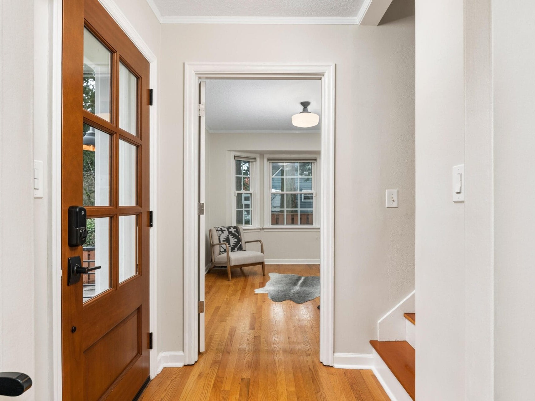 A hallway with wooden floors leads to a bright living room with large windows. On the left, a wooden door with glass panels is partially visible, and on the right, a staircase with wooden steps ascends. A gray rug and an armchair are in the living room.