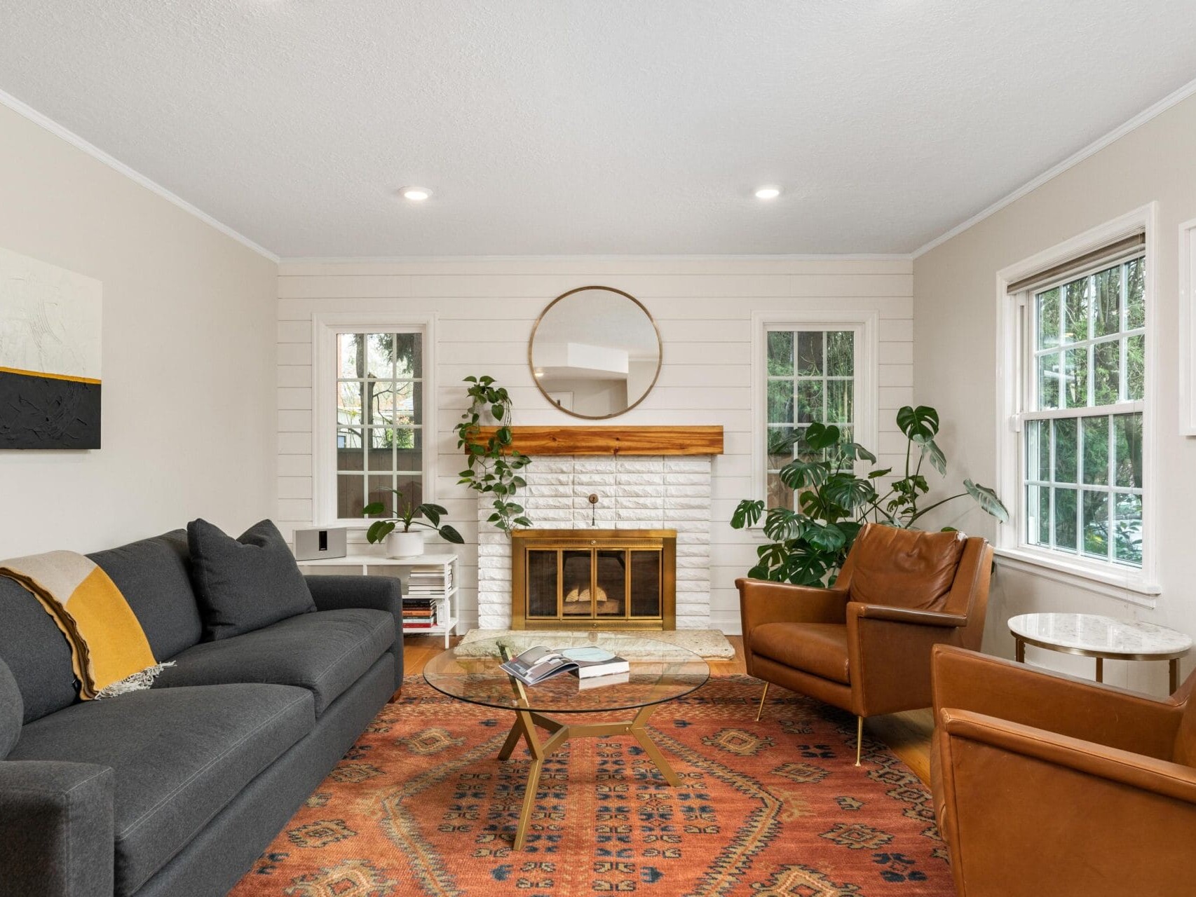 A cozy living room with a dark gray sofa, two brown leather chairs, and a round glass coffee table on a patterned rug. A fireplace with a round mirror above it is flanked by windows with plants. Bright and welcoming atmosphere.