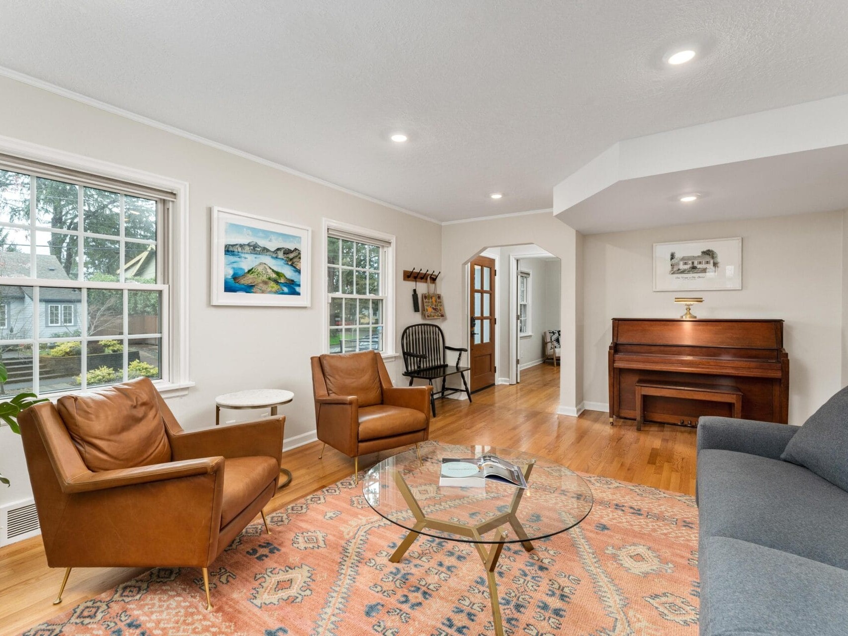 A cozy living room featuring a glass coffee table on a patterned rug, two leather armchairs, a gray sofa, and a wooden piano. The walls are adorned with framed art, and a large window offers natural light.