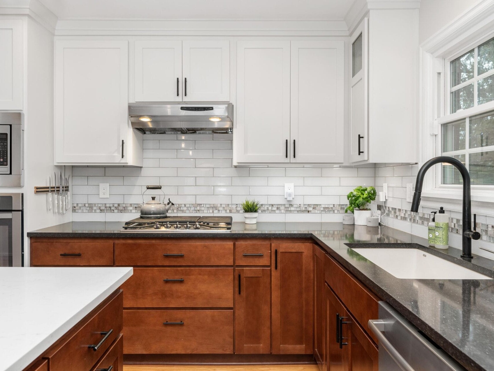 A modern kitchen with white upper cabinets and brown lower cabinets. A stainless steel stove with a kettle sits below a range hood. Theres a gray countertop, a black faucet, and small plants near a window. White subway tiles serve as backsplash.