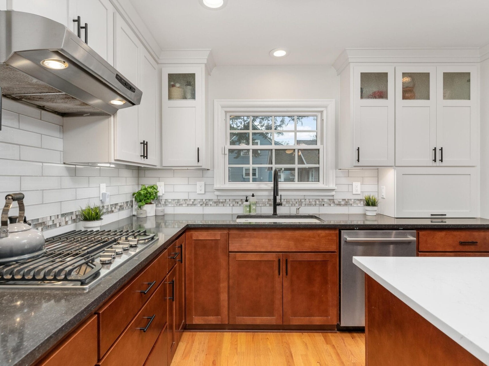 A modern kitchen with white upper cabinets and dark wood lower cabinets. There is a stainless steel stove with a kettle, a dishwasher, and a double-basin sink beneath a window. The countertops are black and white. Small plants decorate the space.