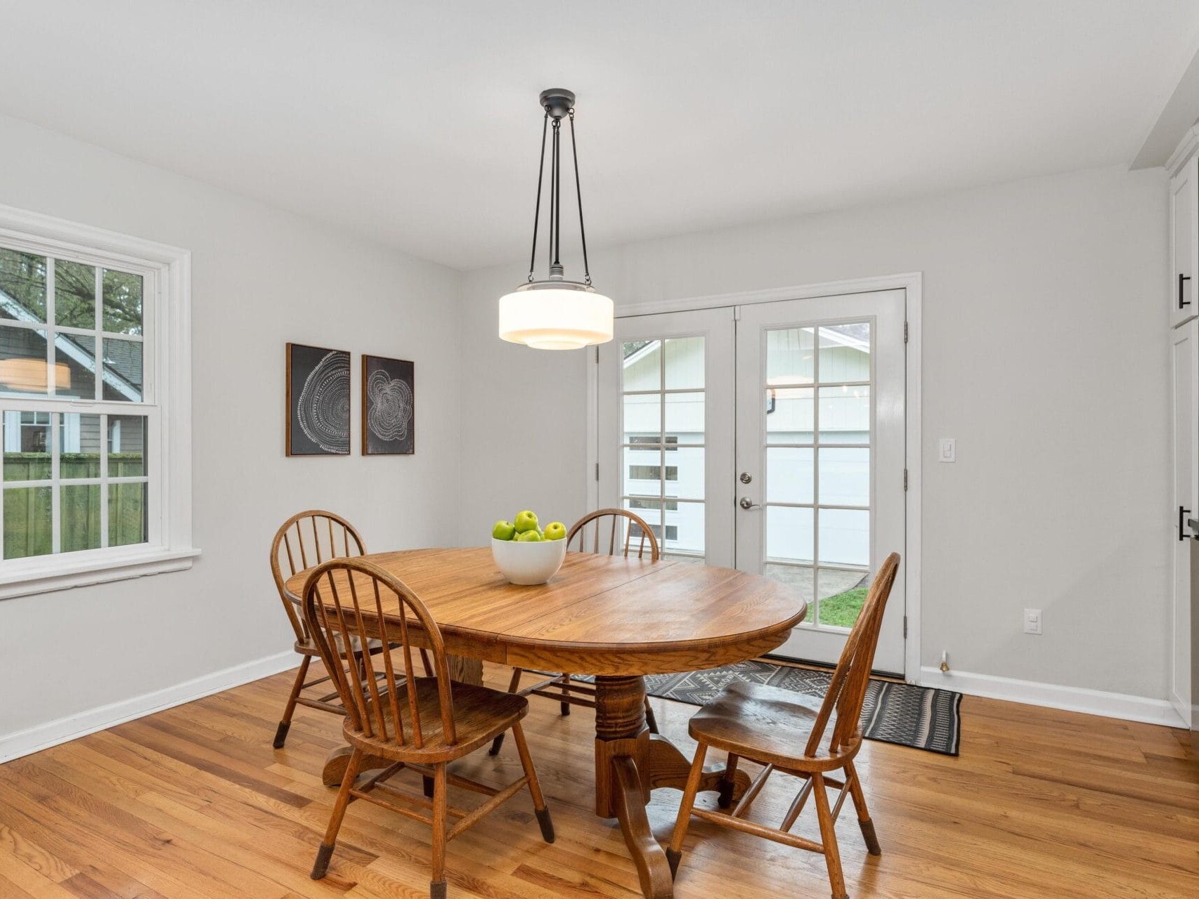 A cozy dining room with a round wooden table and four chairs. A bowl of green apples sits on the table. Two abstract paintings hang on the wall, and a modern pendant light hangs above. French doors and a window let in natural light.