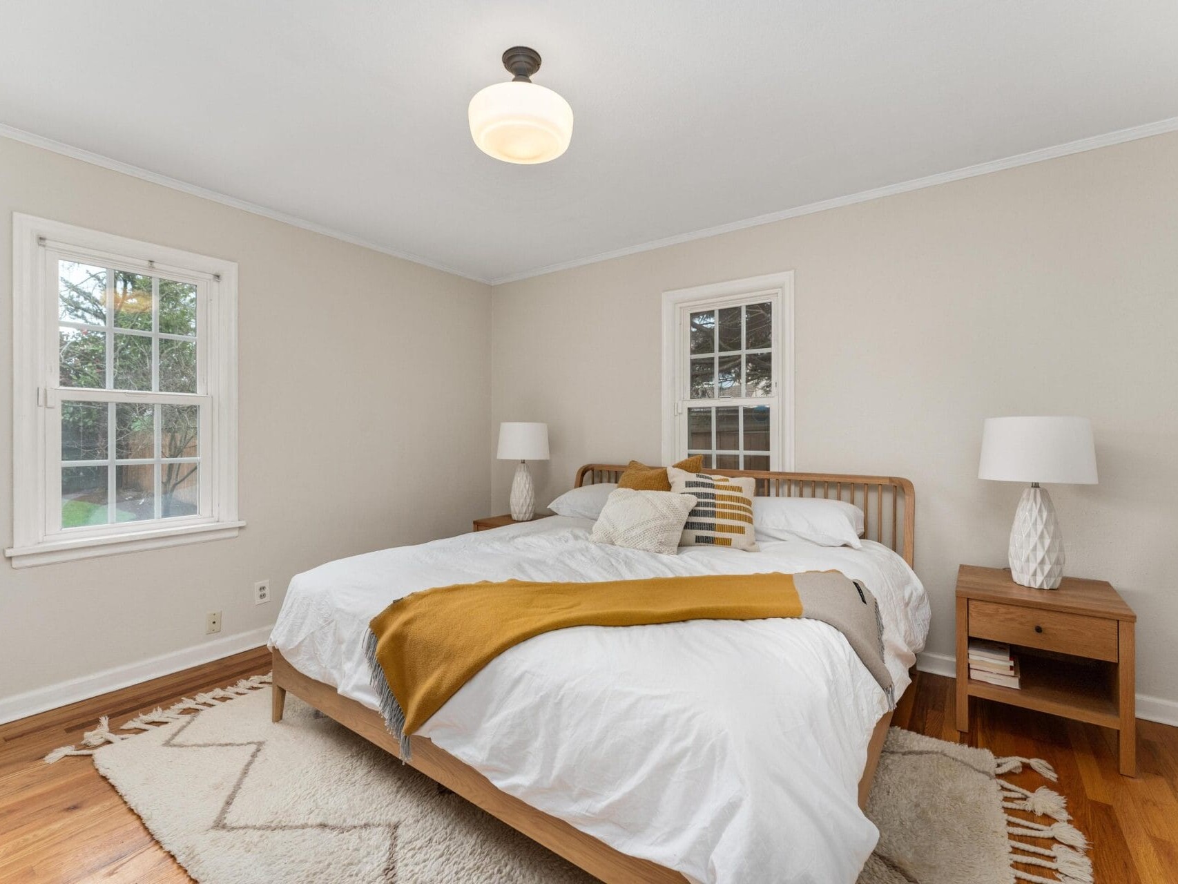 A cozy bedroom with a wooden bed frame featuring white bedding and a yellow throw. Two wooden nightstands, each with a lamp, flank the bed. Natural light enters through two windows, while a geometric rug covers the wooden floor.