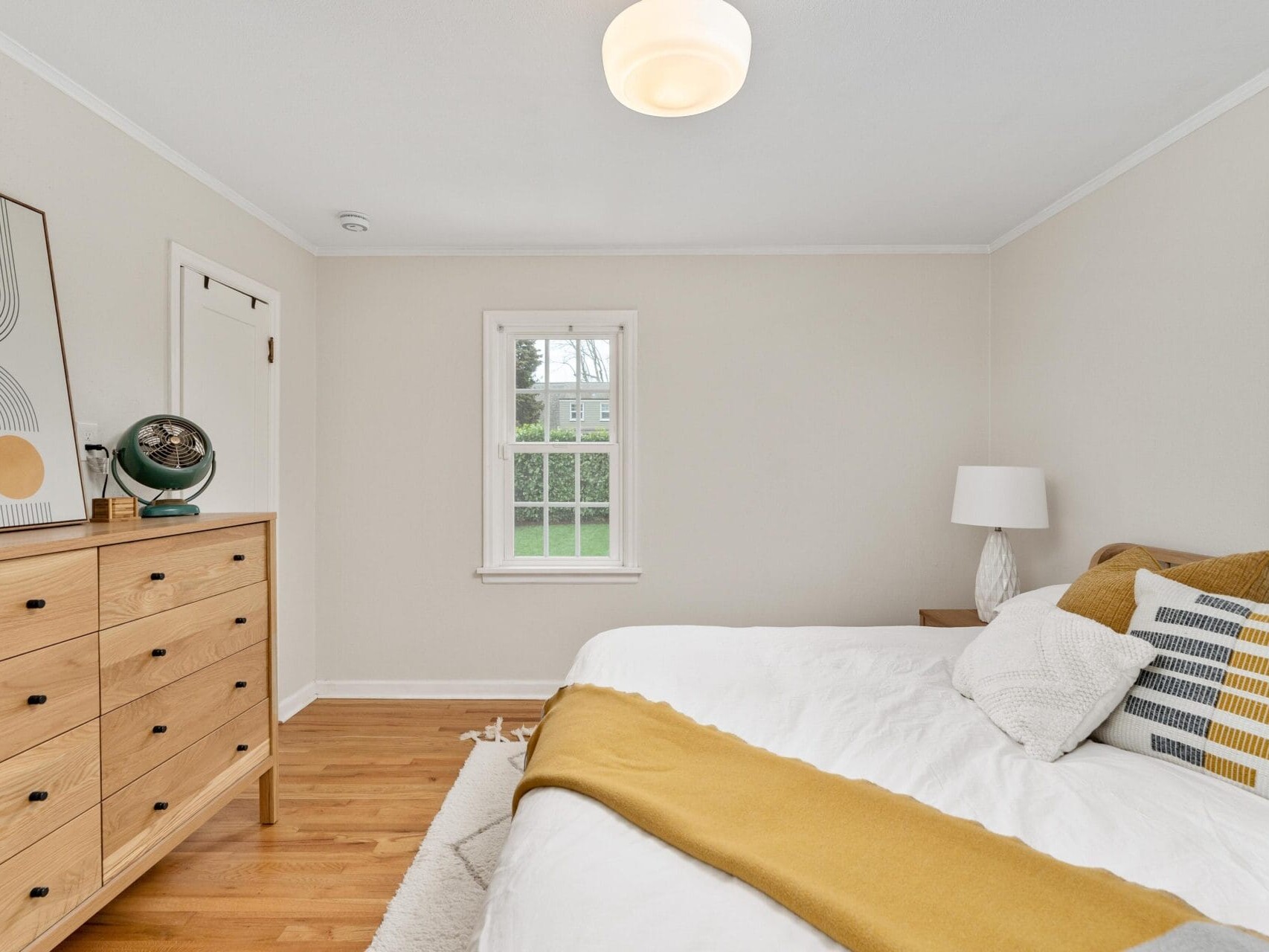 A minimalist bedroom with a white bed, mustard yellow accents, and a wooden dresser. A large abstract art piece leans against the wall. A window overlooks a green yard, and a modern lamp hangs from the ceiling.