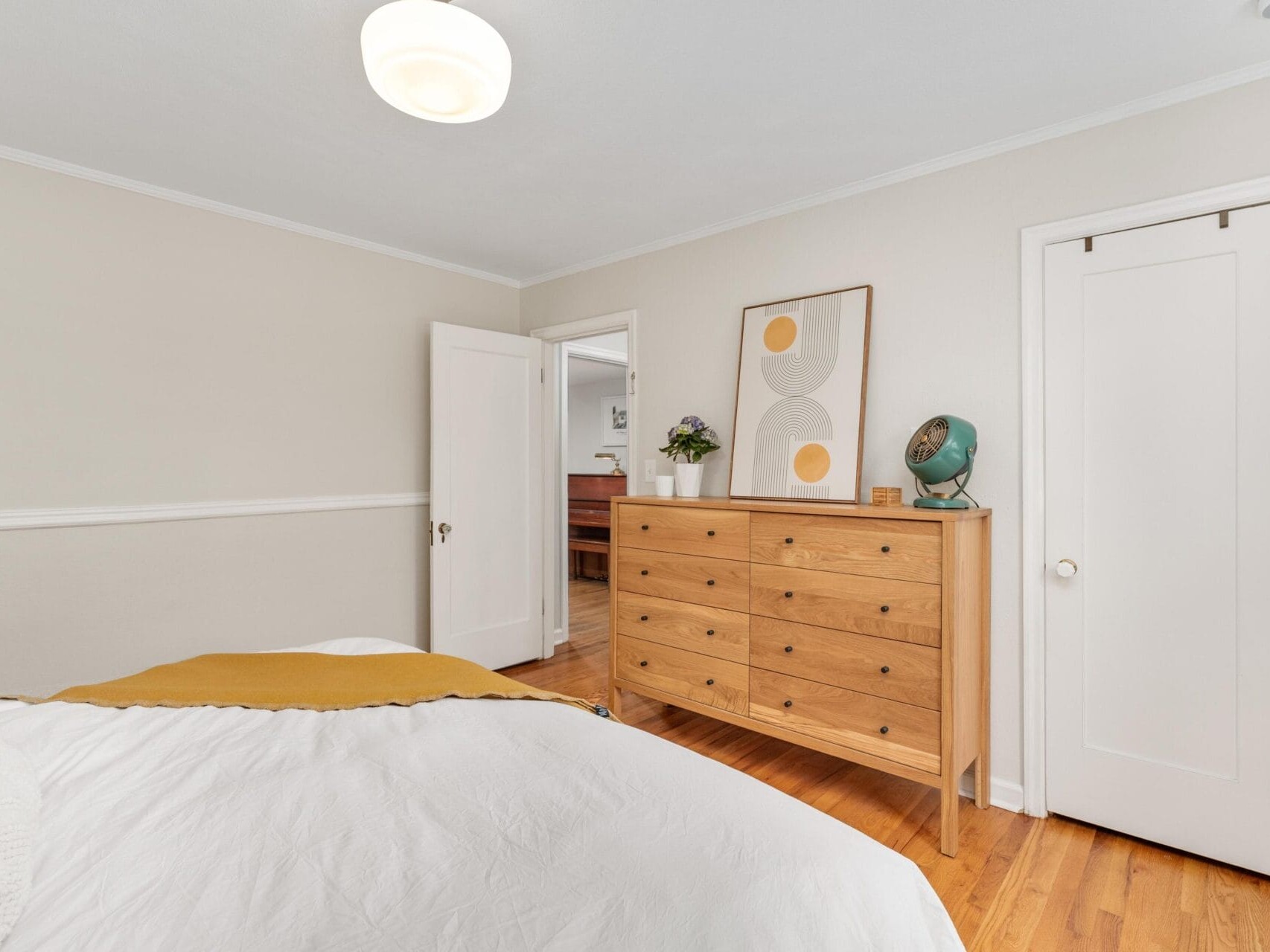 A minimalist bedroom with a white comforter on the bed and a mustard-yellow throw. A wooden dresser against the wall holds a globe and a potted plant. A modern abstract art piece hangs above the dresser. The walls are light beige.