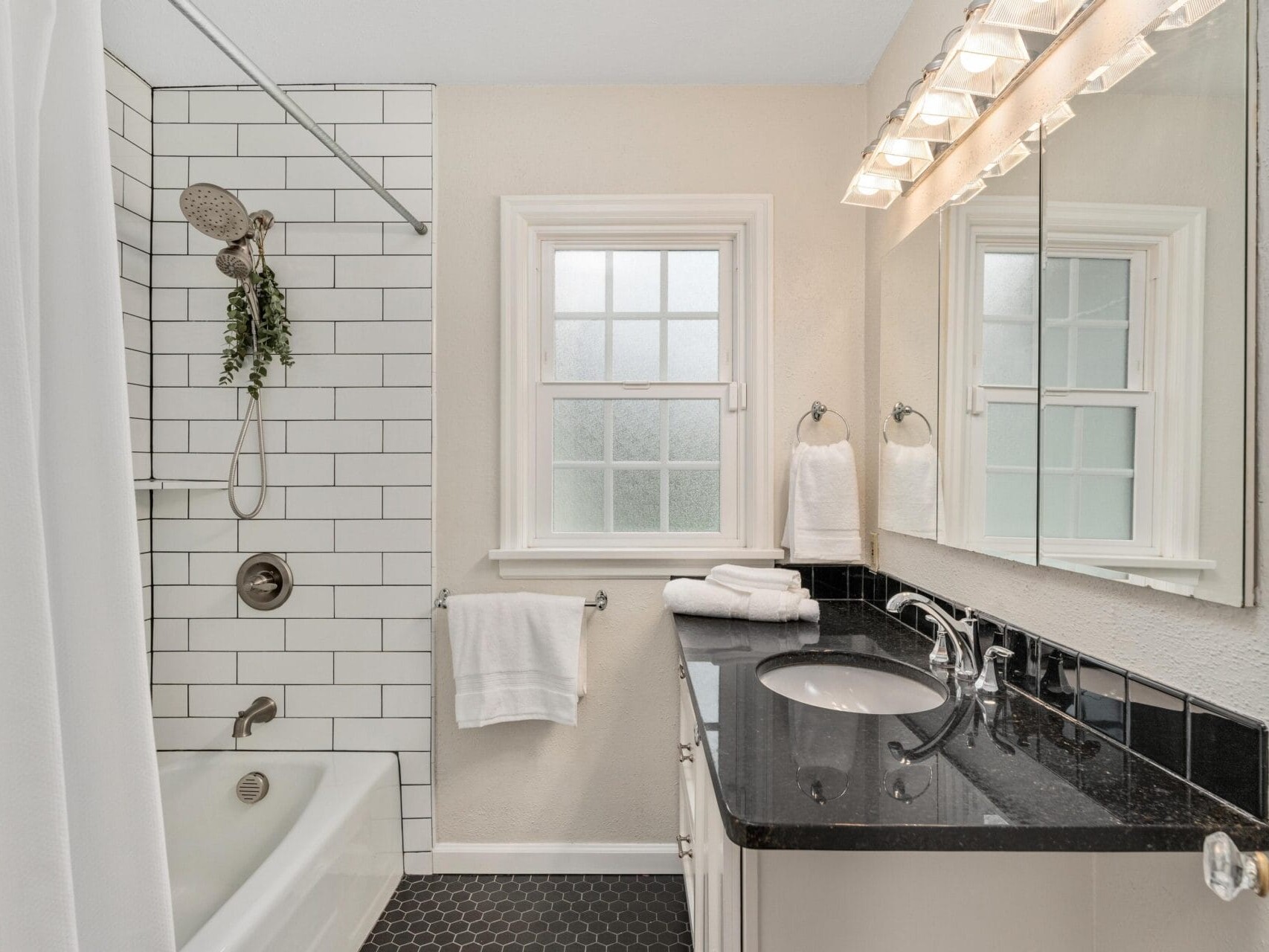 A bathroom with white subway tiles, a bathtub, and a showerhead. A black countertop with a faucet and sink, topped with folded towels, is under a large mirror. The floor has hexagonal tiles, and a window brings in natural light.