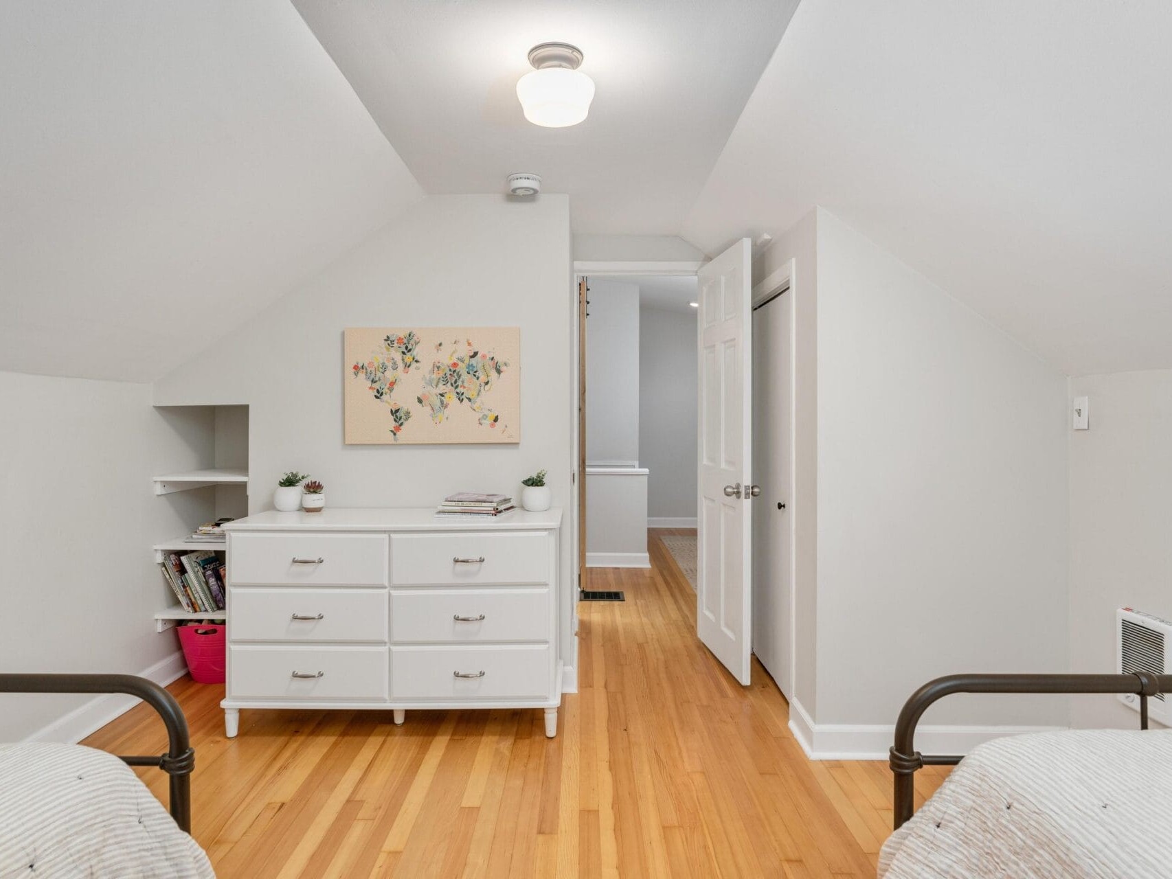 A minimalist bedroom with two twin beds, a white dresser between them, and light wood flooring. Theres artwork above the dresser and a small bookshelf on the left. A hallway is visible in the background with a door to the right.