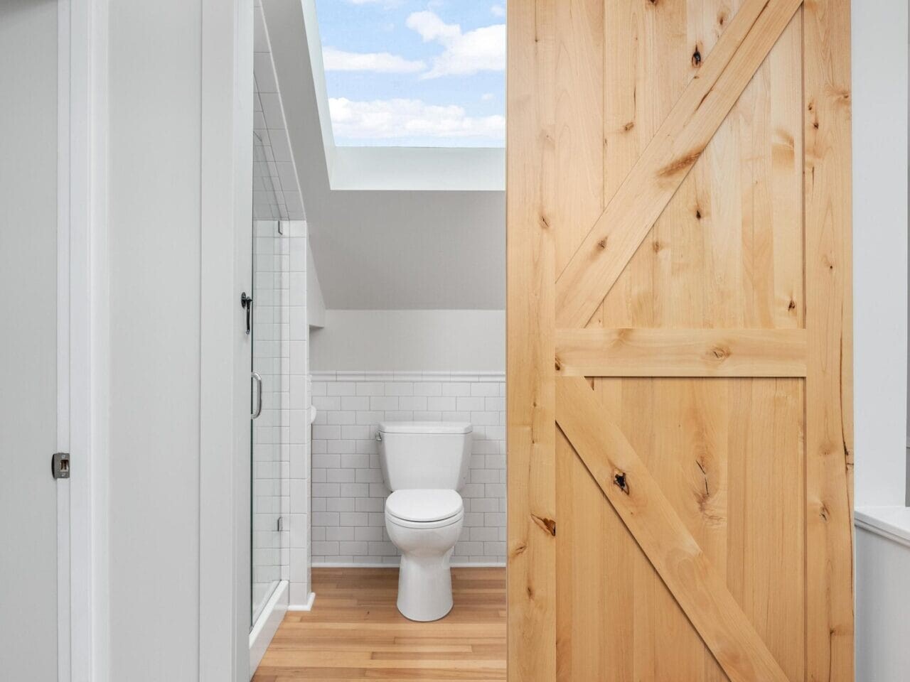A bathroom with a sliding wooden barn door partially open. Inside, theres a white toilet and a shower with white subway tiles. The floor is light wood, and theres a skylight above, revealing a blue sky with clouds.