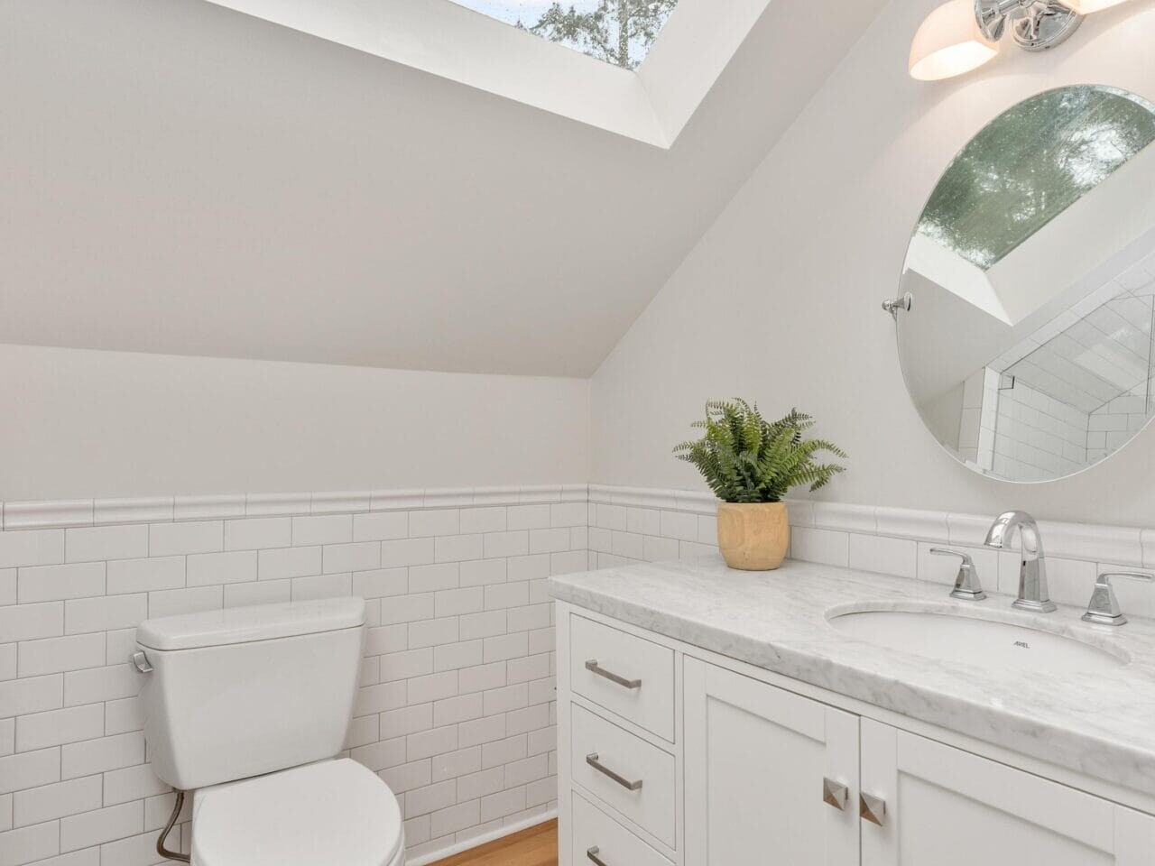 A bright bathroom with a slanted ceiling and skylight. Features include a white vanity with a marble countertop, round mirror, potted plant, modern faucet, and toilet. The walls have white subway tiles, and the floor is hardwood. Sky and trees visible.