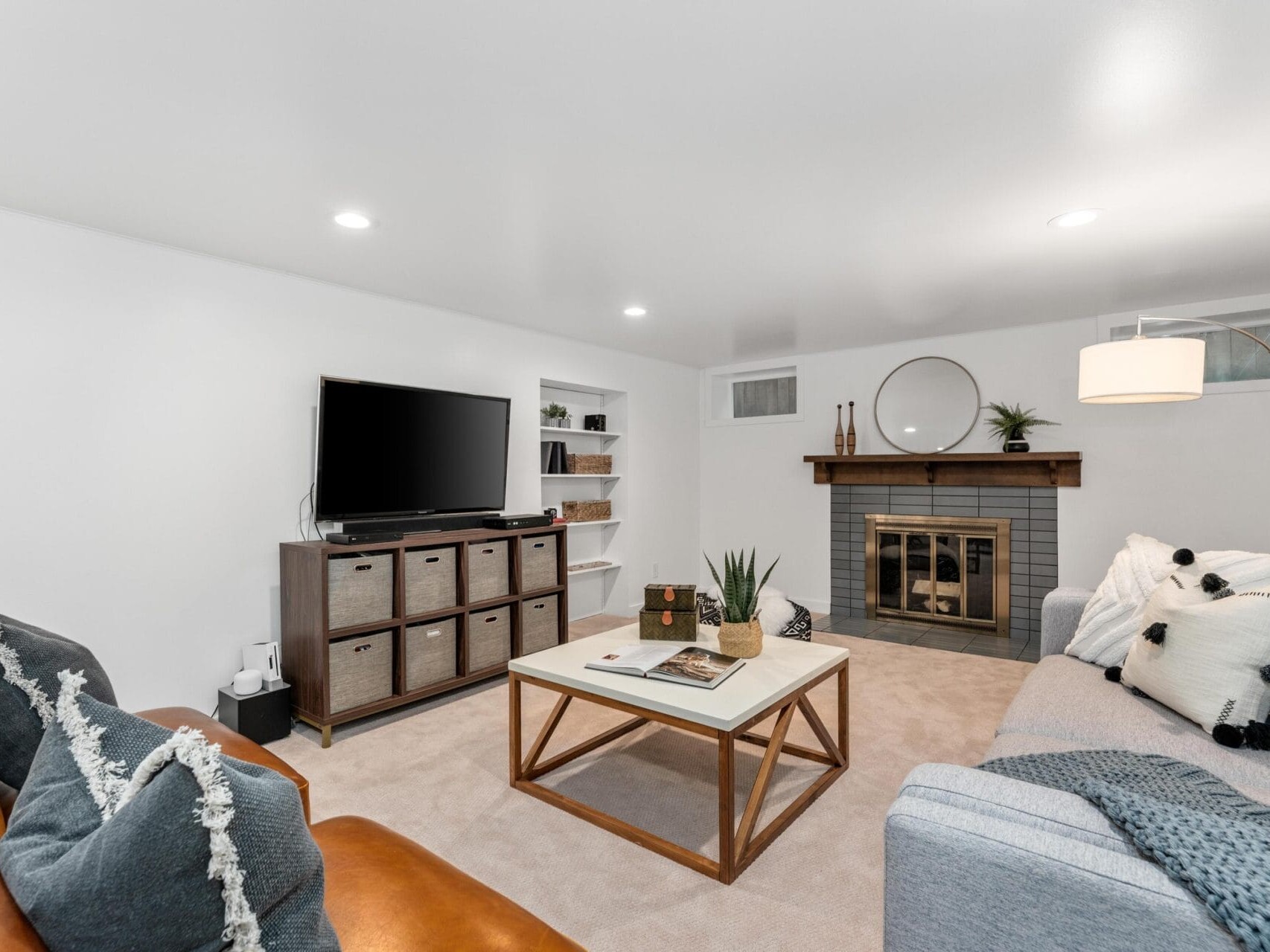 A cozy living room with a gray couch, a brown leather chair, and a large TV on a cabinet. Theres a coffee table in the center and a fireplace with a round mirror above it. Shelves with decor and a floor lamp complete the space.