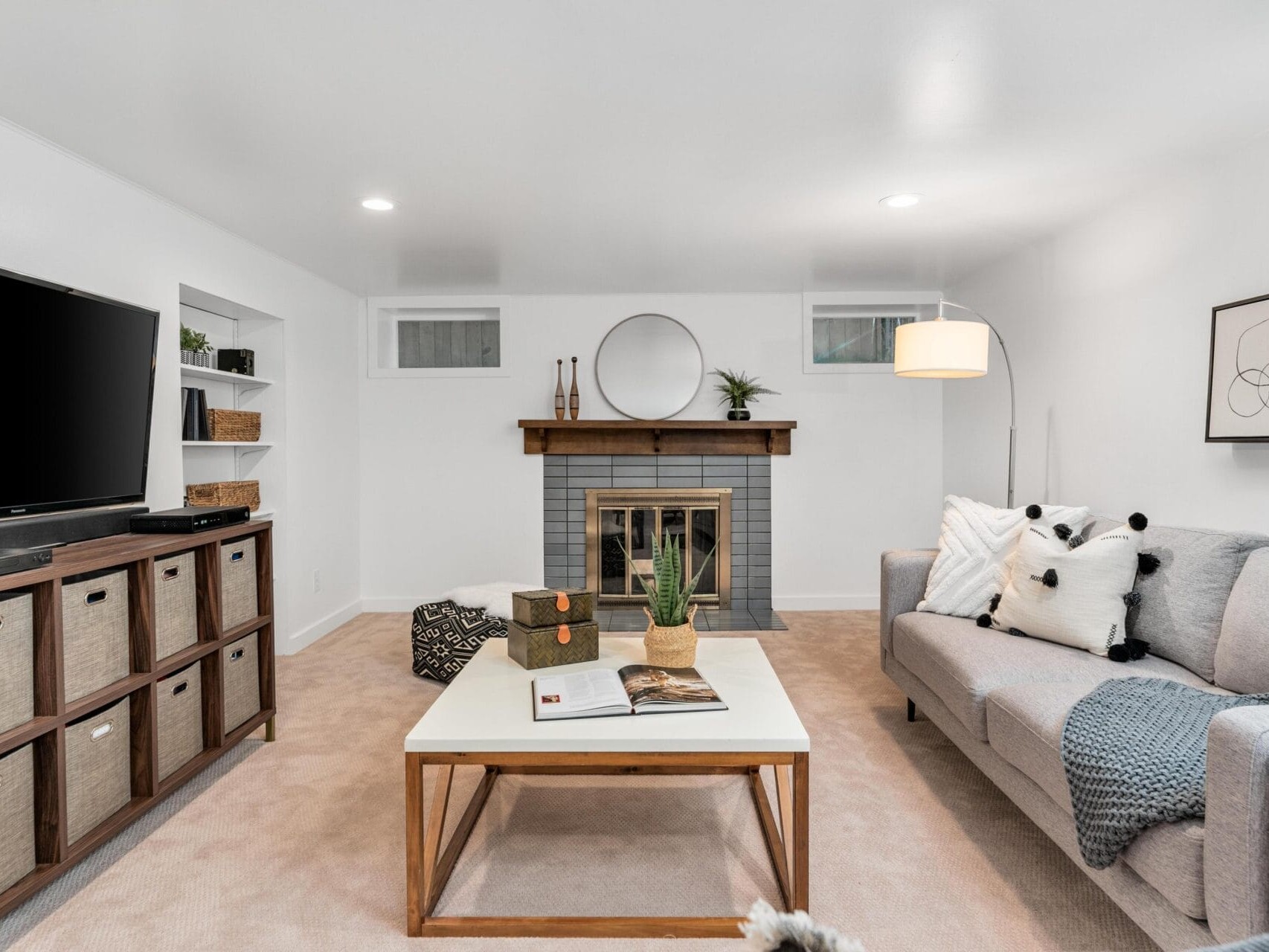 A cozy living room with a modern gray sofa adorned with black and white pillows, a coffee table with books, a TV on a stand with storage baskets, a brick fireplace with a round mirror above, and a tall floor lamp in the corner.