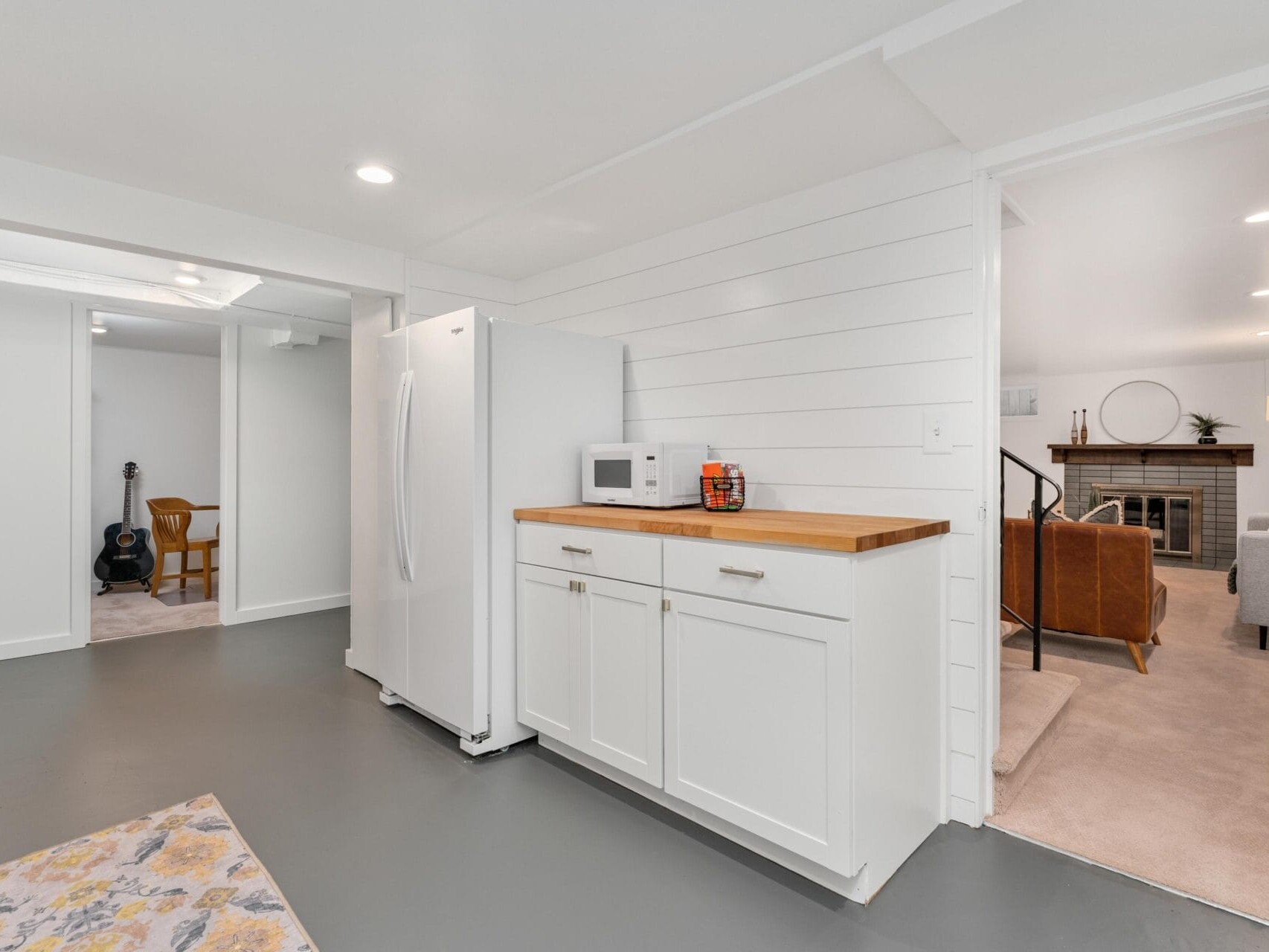 A modern basement features white shiplap walls and a polished gray floor. A white refrigerator and cabinet with a wooden countertop hold a microwave and decor. An adjoining carpeted living area with a fireplace and a guitar in the corner are visible.