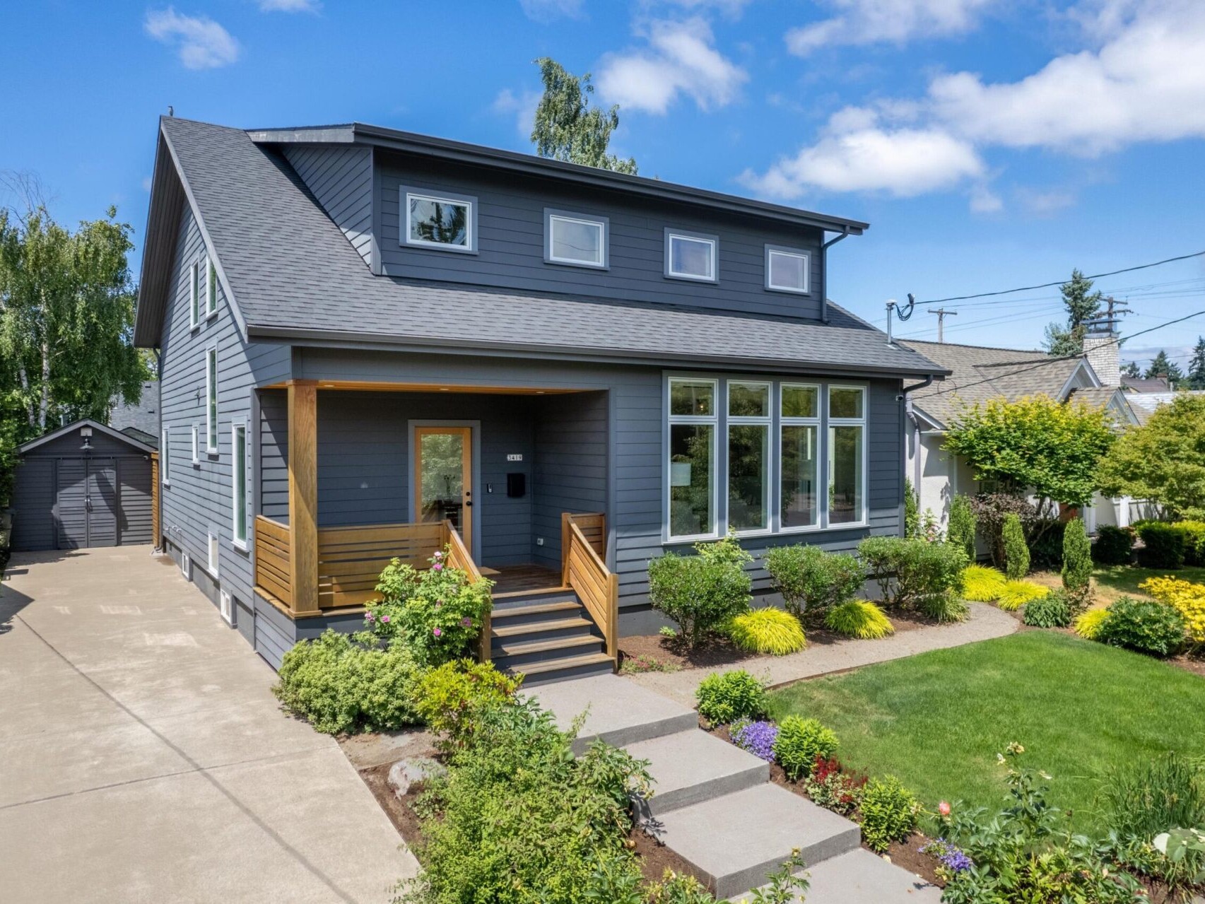 A modern blue-gray two-story house with a well-maintained front yard. The entrance features a small porch with wooden railings, surrounded by vibrant green bushes and colorful flowers. A driveway leads to a detached garage in the background.
