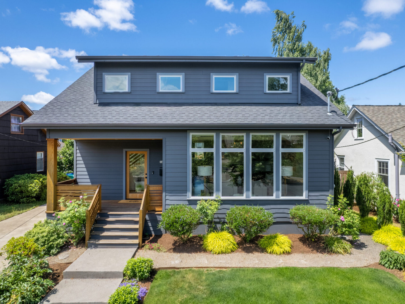 A modern two-story house with a gray exterior, large windows, and a wooden porch. It's surrounded by a well-maintained garden with green shrubs and a lawn. The sky is clear with a few clouds—a perfect gem in the Portland Oregon real estate market.