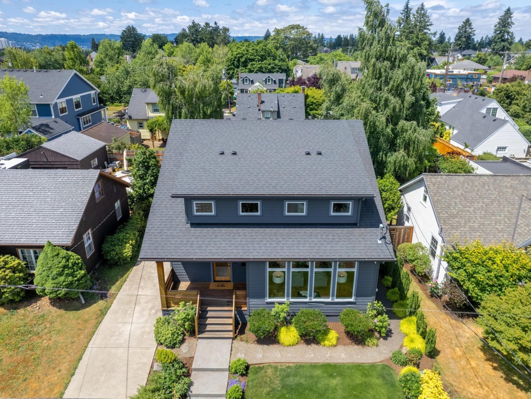 An aerial view of a suburban neighborhood with a prominent two-story gray house featuring a well-maintained garden and a large front window. The house is surrounded by similar homes, trees, and greenery under a partly cloudy sky.
