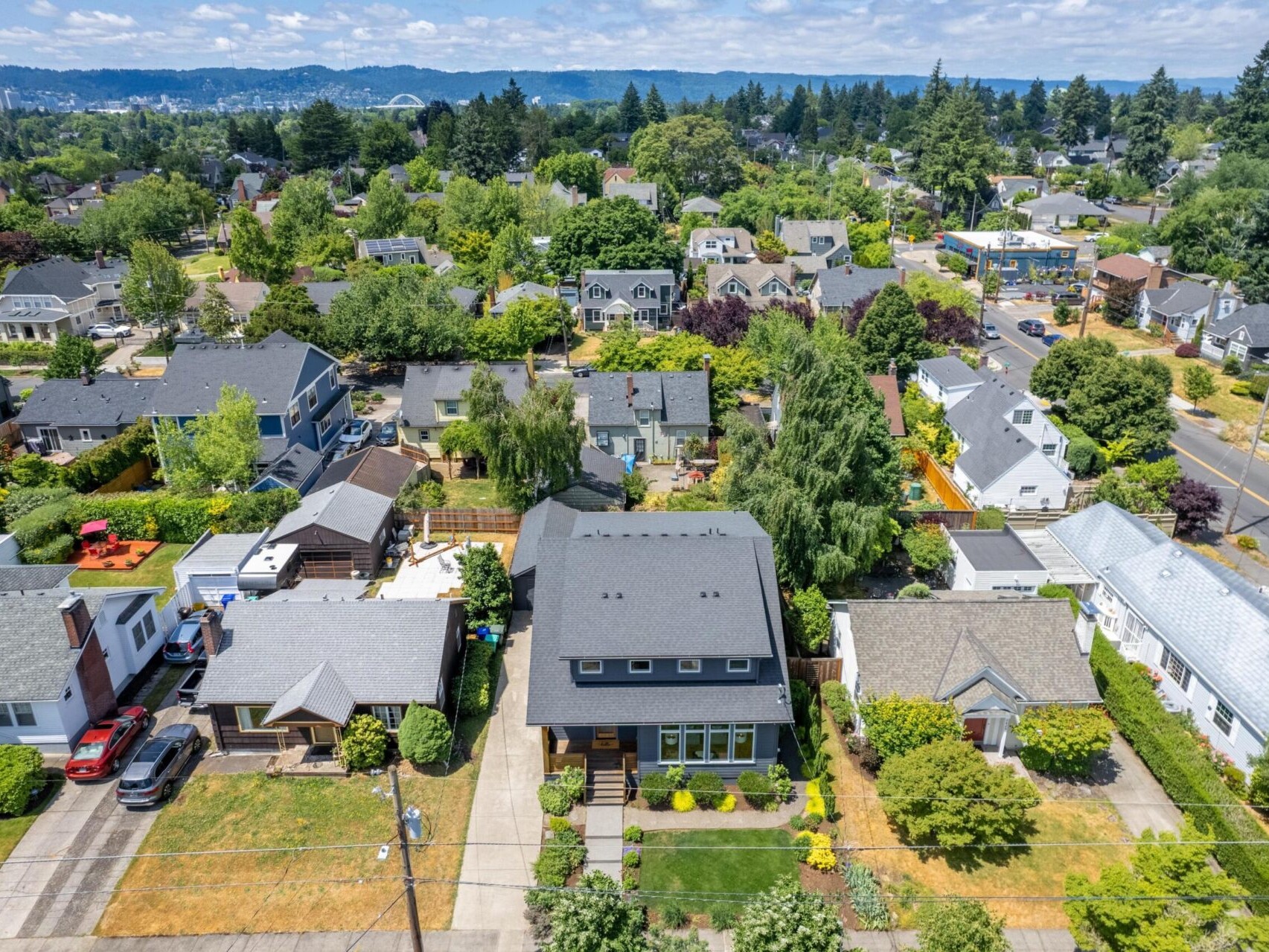 An aerial view of a suburban neighborhood with modern houses, lush green trees, and well-maintained lawns. A main road runs alongside, with hills and a bridge visible in the background under a partly cloudy sky.