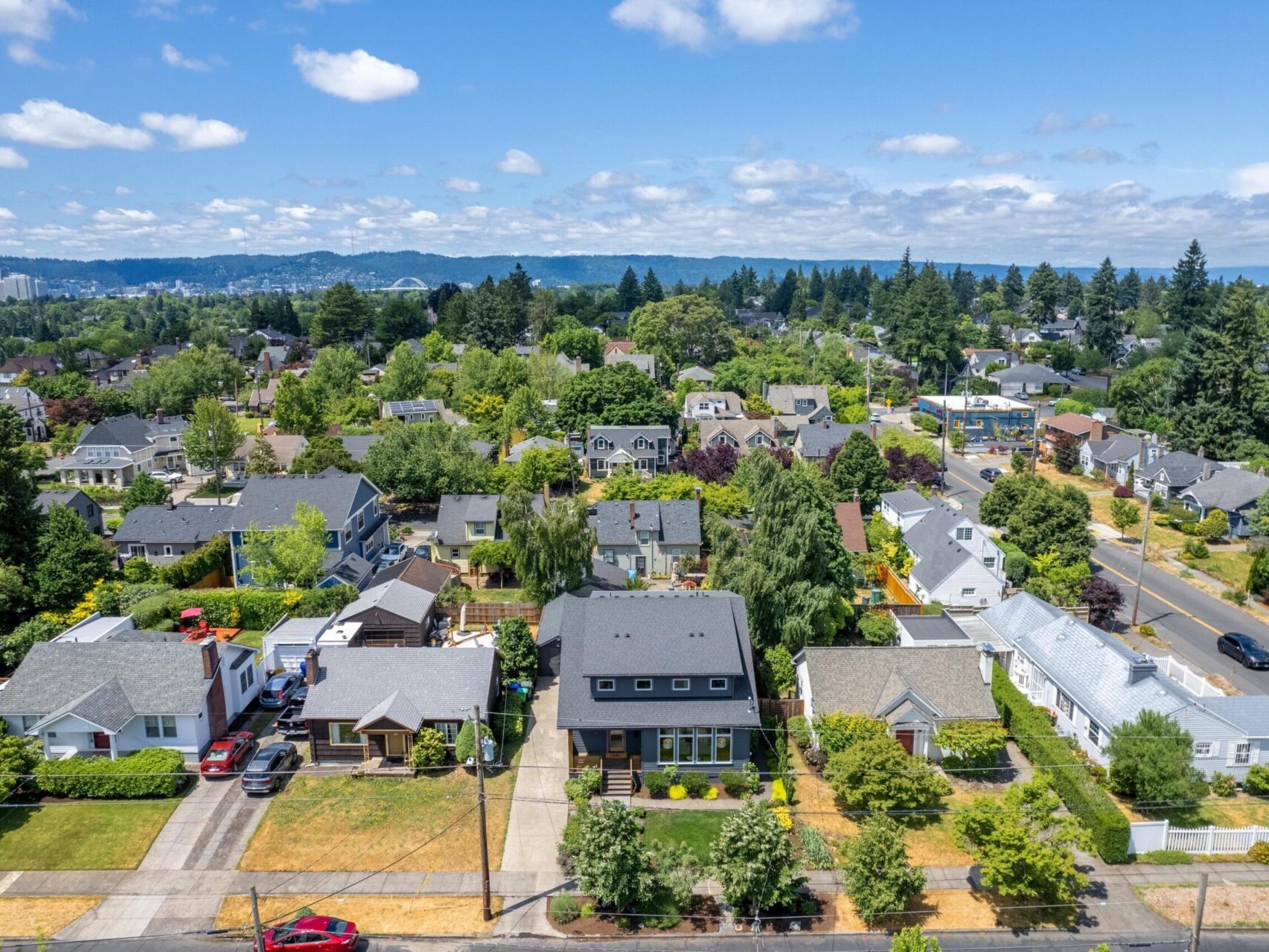 Aerial view of a suburban neighborhood with tree-lined streets and houses. The area is surrounded by greenery under a bright blue sky with scattered clouds. A distant cityscape and hills are visible on the horizon.