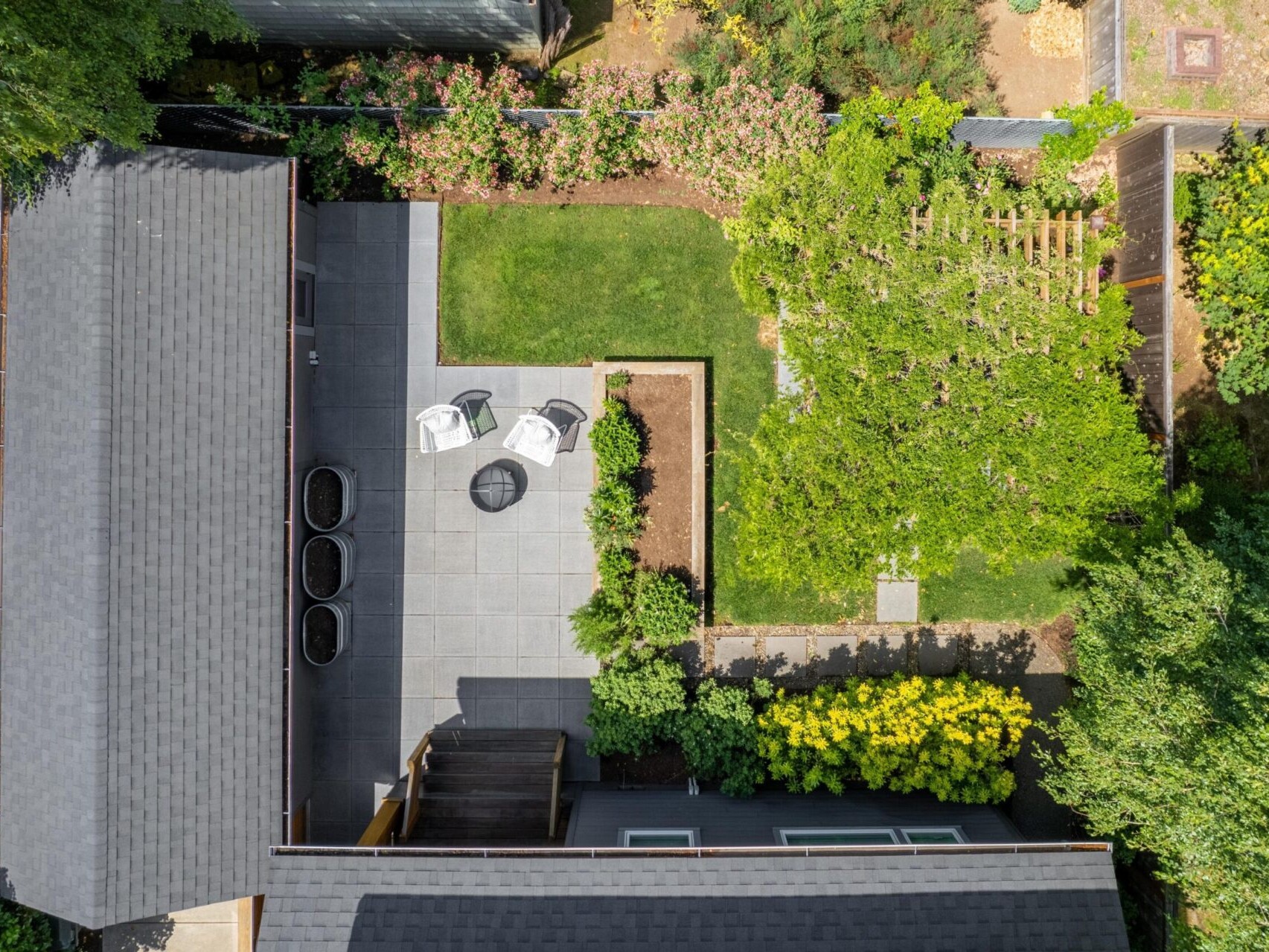 Aerial view of a backyard with a patio featuring two white chairs and a small table on a tiled area. Surrounding greenery includes trees, shrubs, and neatly trimmed grass. A nearby pergola is partially visible beneath a canopy of foliage.