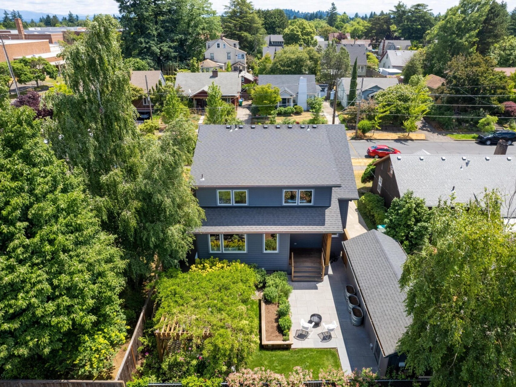 Aerial view of a two-story house surrounded by trees in a suburban neighborhood. The house has a dark roof and a well-maintained yard with a fenced garden and a patio set. Other homes and tree-lined streets are visible in the background.