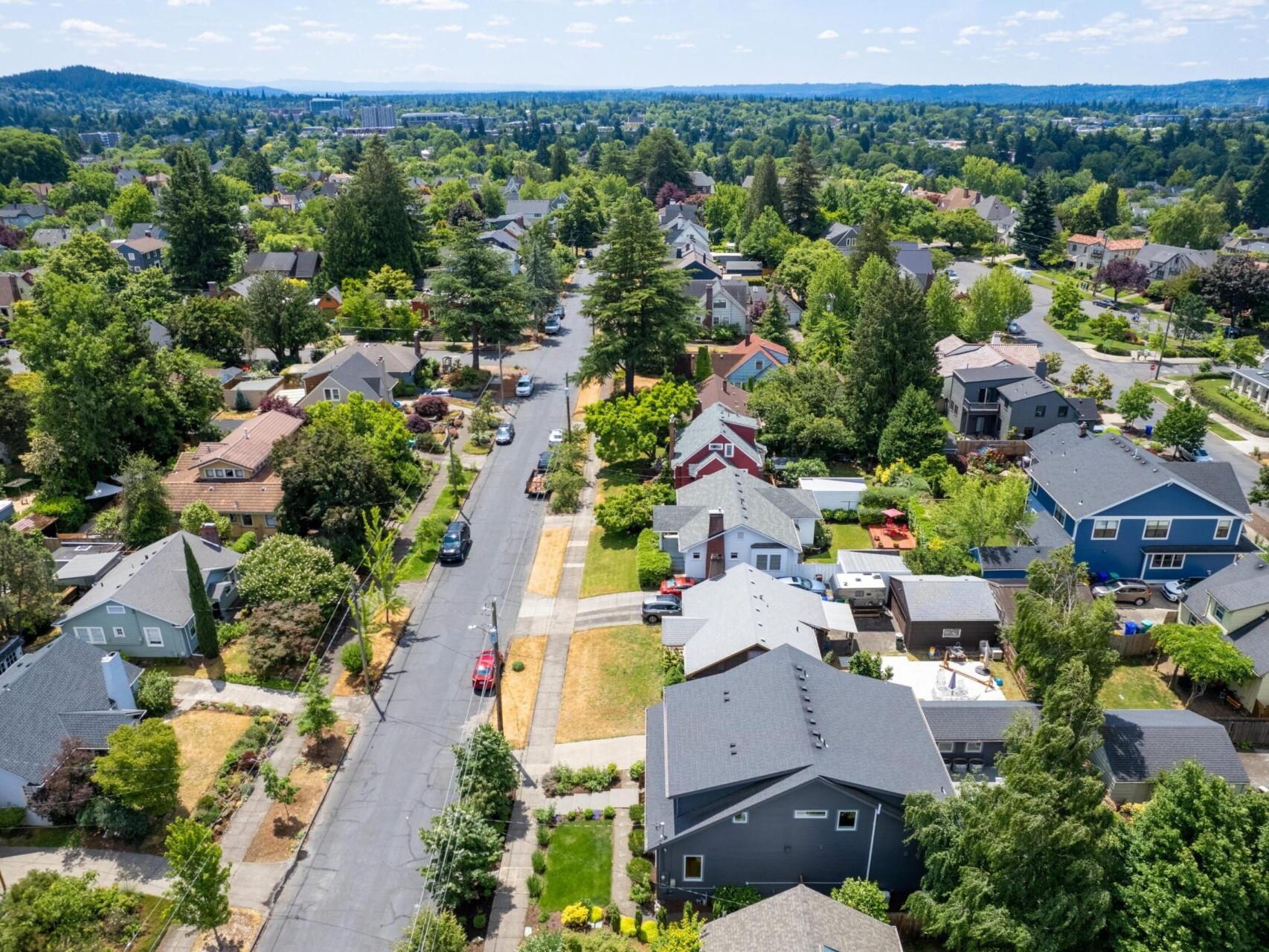 Aerial view of a suburban neighborhood with tree-lined streets and houses. The area features a mix of homes, lush greenery, and a clear blue sky, creating a peaceful residential setting.