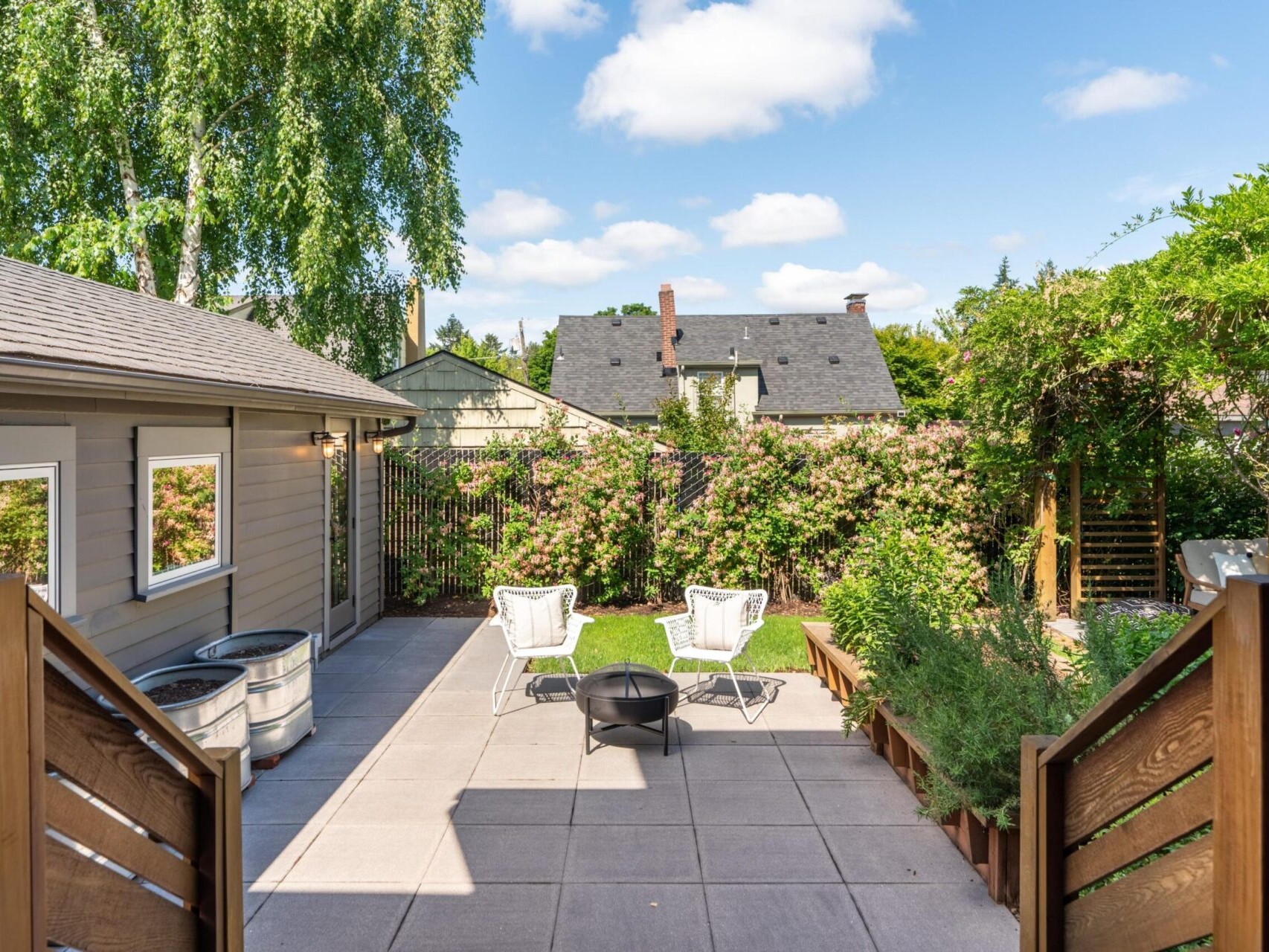 A spacious backyard with a concrete patio features two white chairs and a black fire pit. The area is surrounded by lush greenery and a wooden fence. A house with a gray exterior and a tree in the background is visible under a clear blue sky.