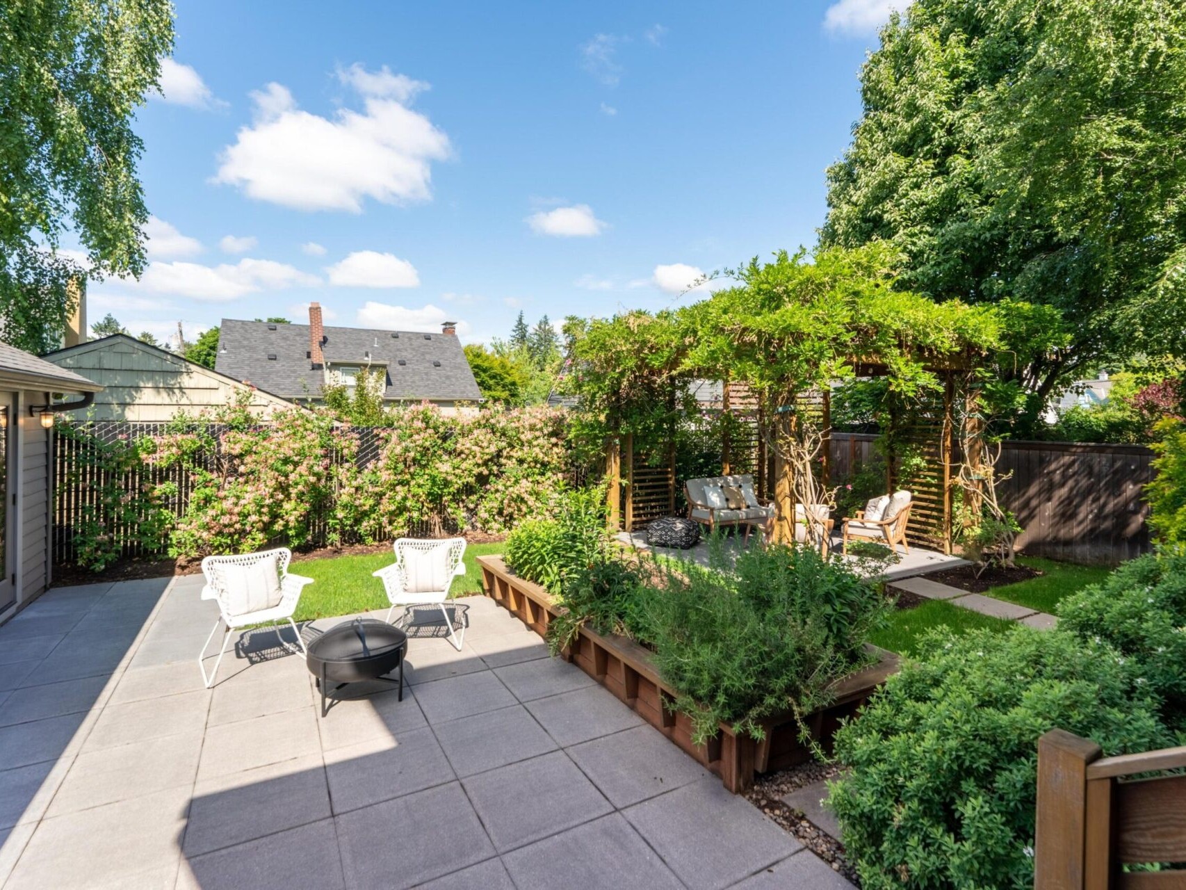 A sunny backyard with a patio featuring white chairs and a fire pit, surrounded by lush greenery. A wooden pergola with seating is to the right, near a tall tree and shrubs. Houses with sloped roofs are visible in the background.