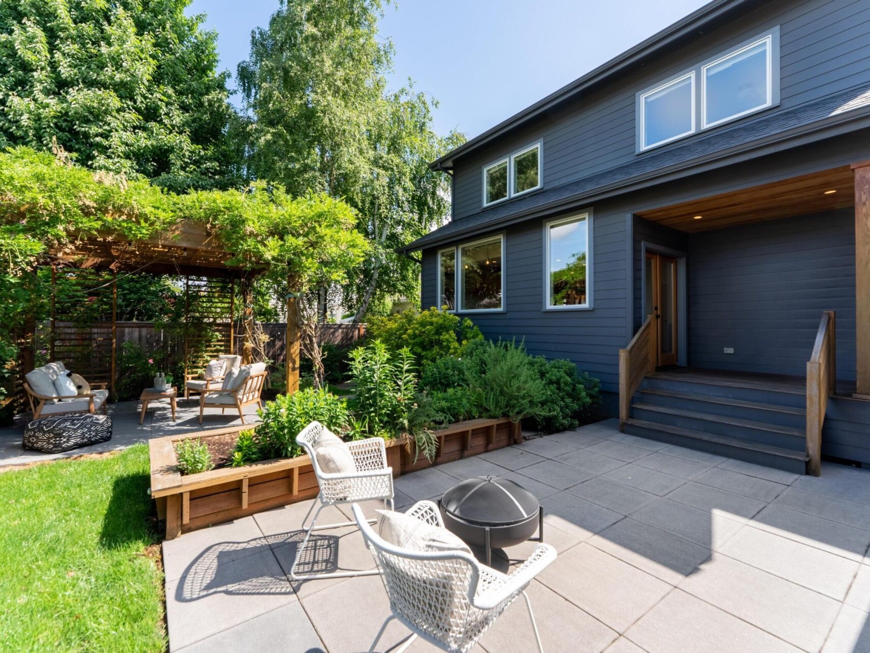 A backyard with a modern gray house, featuring a patio. The area includes white chairs and a small table on a tiled surface. On the left, a cozy seating area is shaded by a wooden pergola with greenery. Lush trees and plants surround the space.
