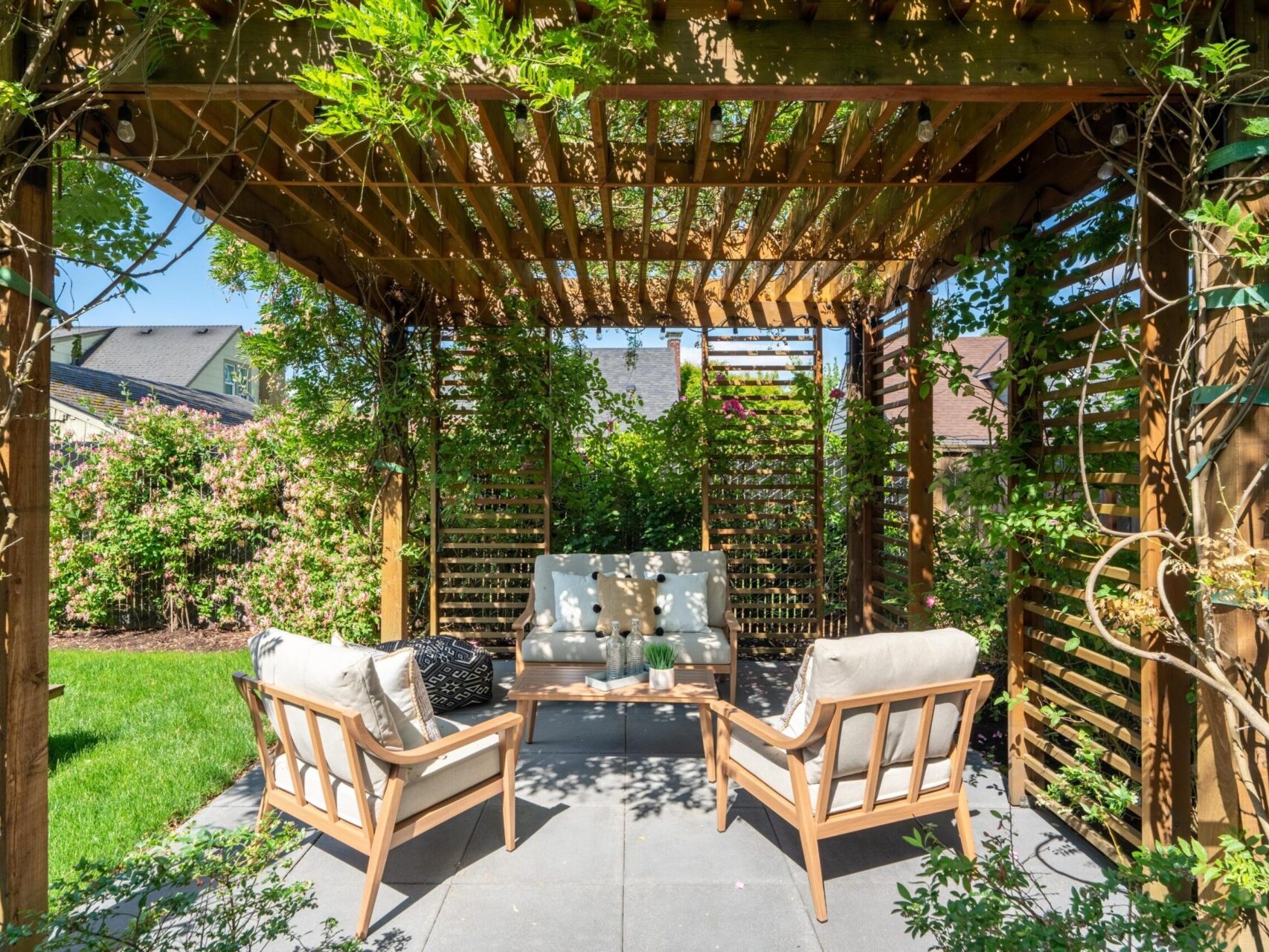 A wooden pergola with climbing plants provides shade over a cozy patio area featuring cushioned chairs and a coffee table. Sunlight filters through the lattice roof, and the surrounding garden is lush with greenery and flowering plants.