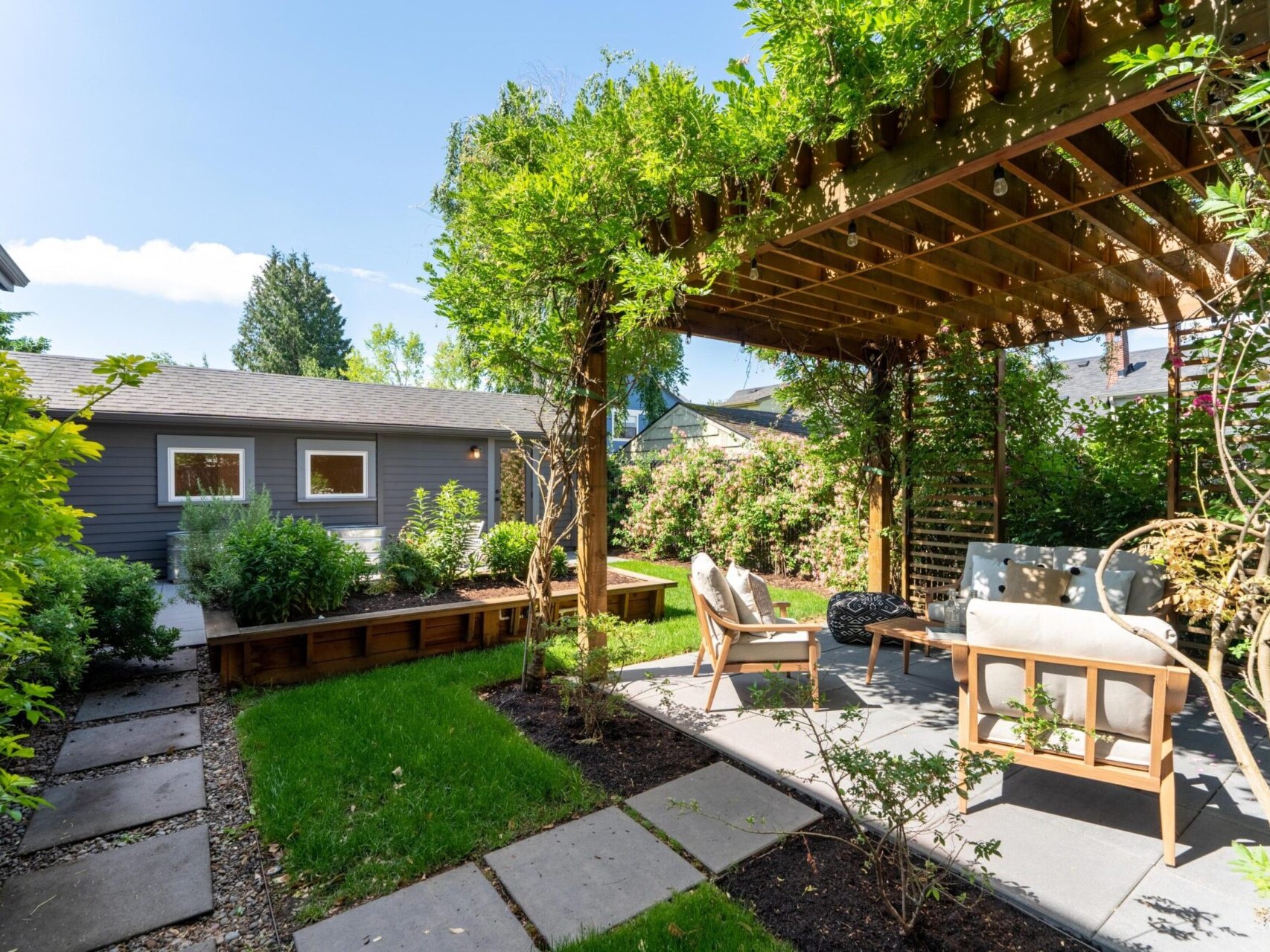 A backyard with a wooden pergola covered in greenery, providing shade over two cushioned chairs and a table on a stone patio. The area is surrounded by lush plants and leads to a small building with a window and gravel pathway.