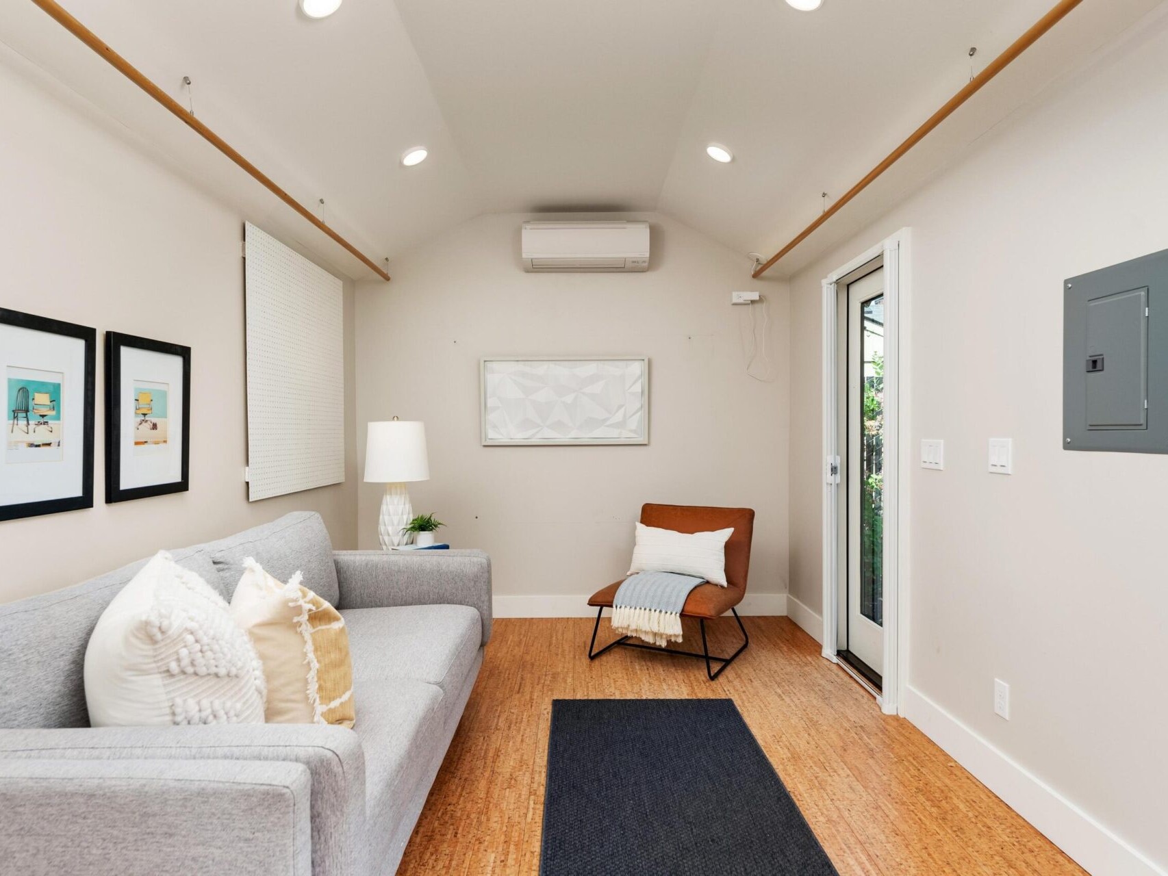 A cozy, modern living room with a gray sofa, two framed artworks on the wall, a small table with a lamp, and an orange accent chair with a blanket. The room has light walls, wooden flooring, a door on the right, and a ceiling air conditioner.
