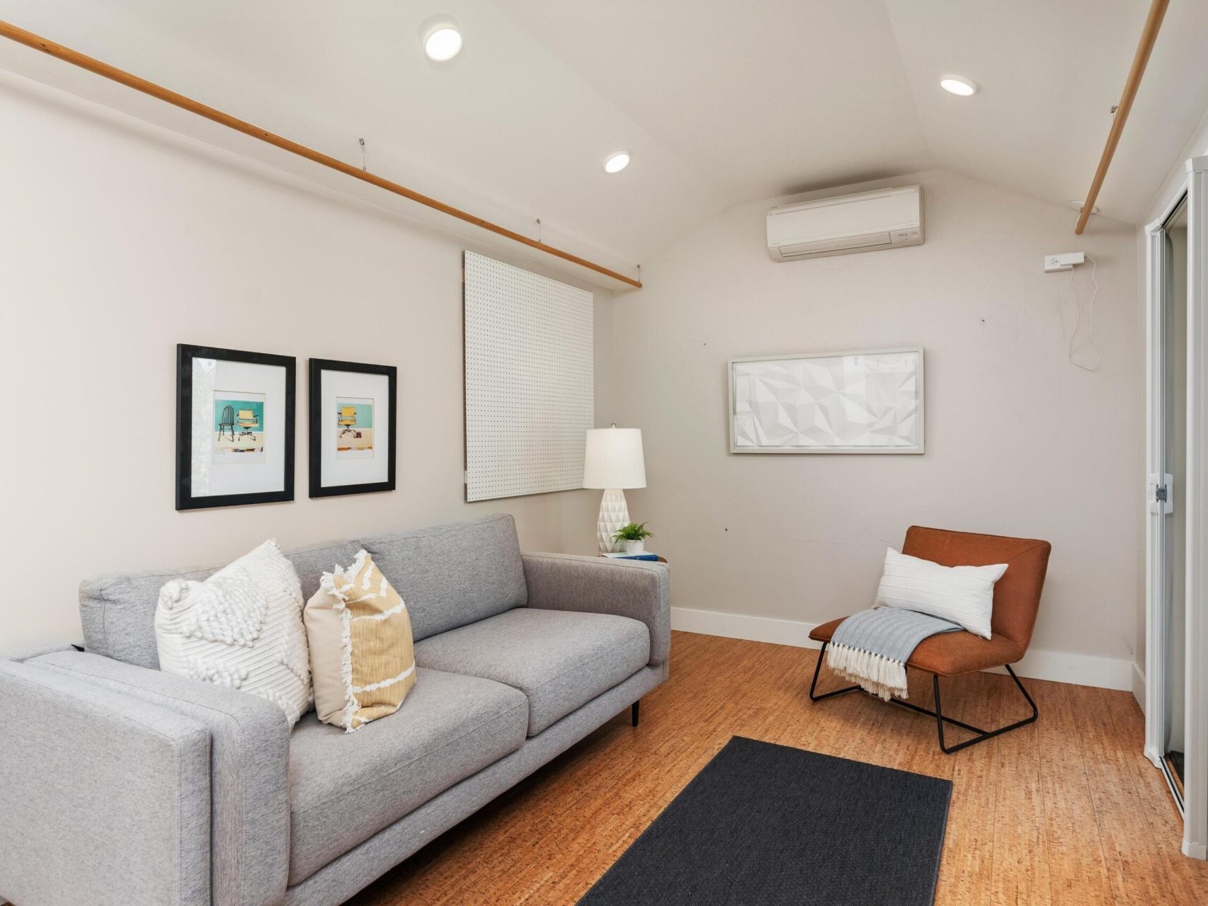 A cozy living room with a gray sofa decorated with cushions, a brown chair with a white throw, and a small lamp on a side table. Two framed artworks and a rectangular piece hang on the white walls. A rolled-up blind covers the window.