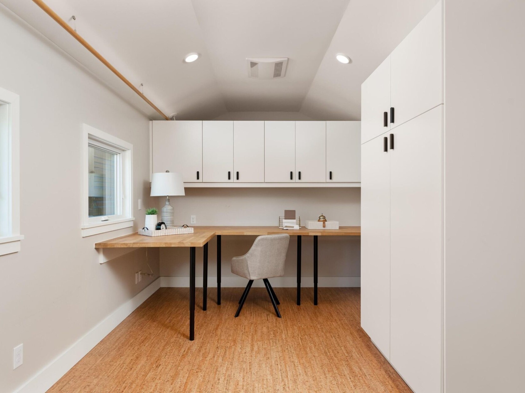 A minimalist home office with a wooden desk, beige swivel chair, white cabinets, and a desk lamp. The room has light-colored walls and a window on the left, allowing natural light in. The floor is covered in wood-like material.