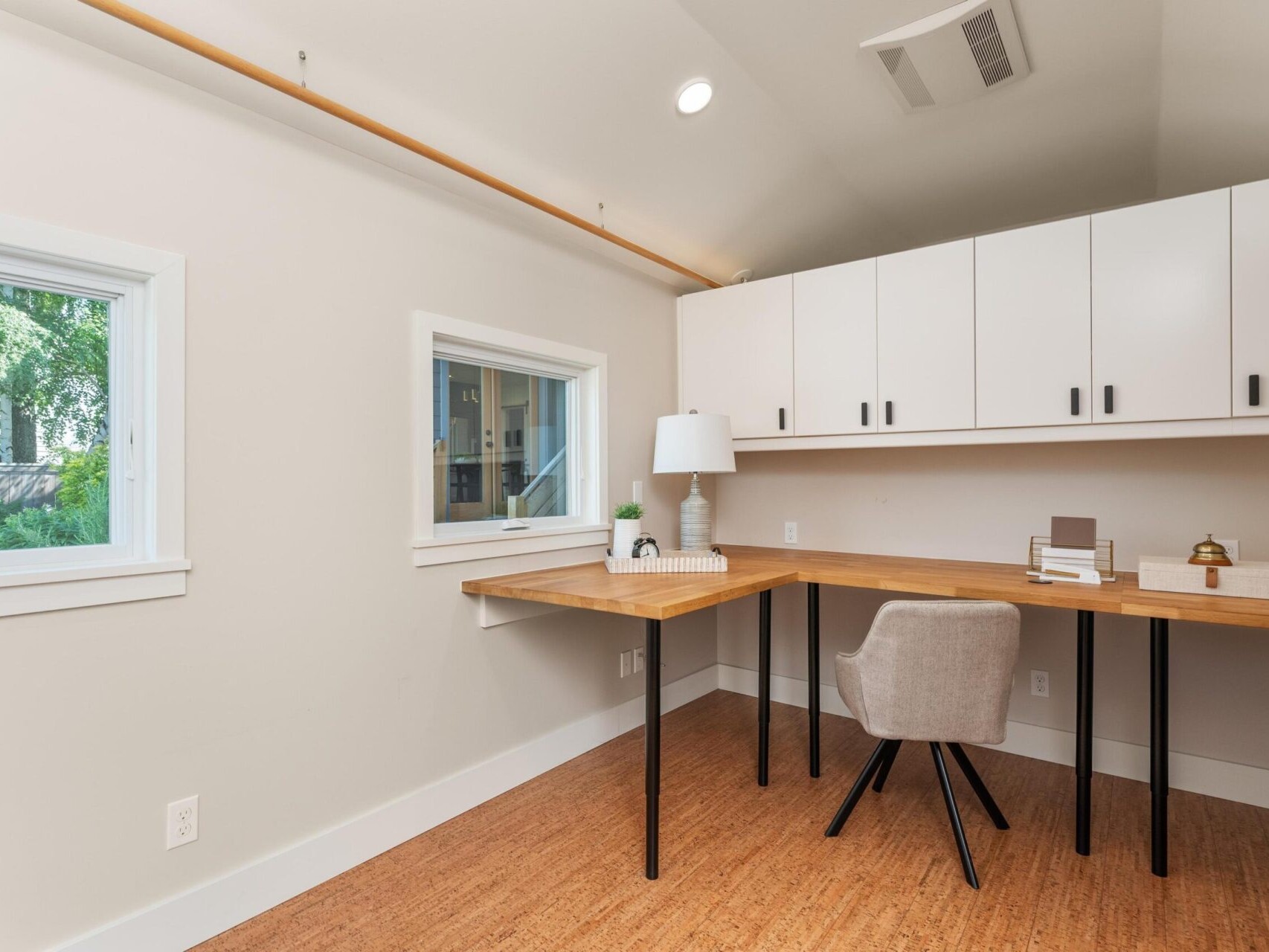 A minimalist home office featuring a corner wooden desk with a gray swivel chair, white overhead cabinets, and a table lamp. Large windows provide natural light, while the light-colored walls and flooring create an airy atmosphere.
