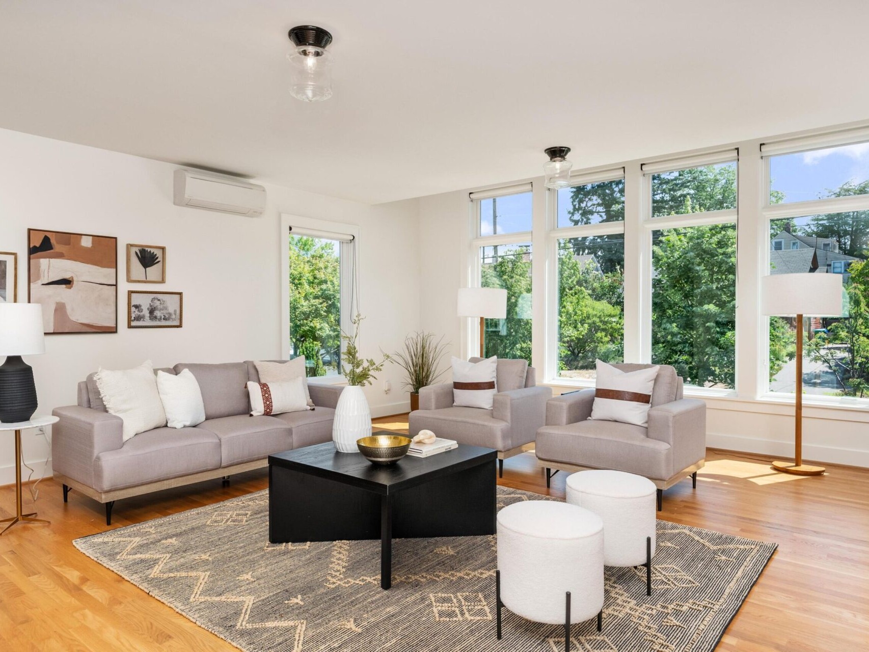 A modern living room with two gray sofas, armchairs, and white poufs. The room features a black coffee table, a patterned rug, and large windows offering a view of greenery. Minimalist decor and artwork adorn the walls, with ample natural light.