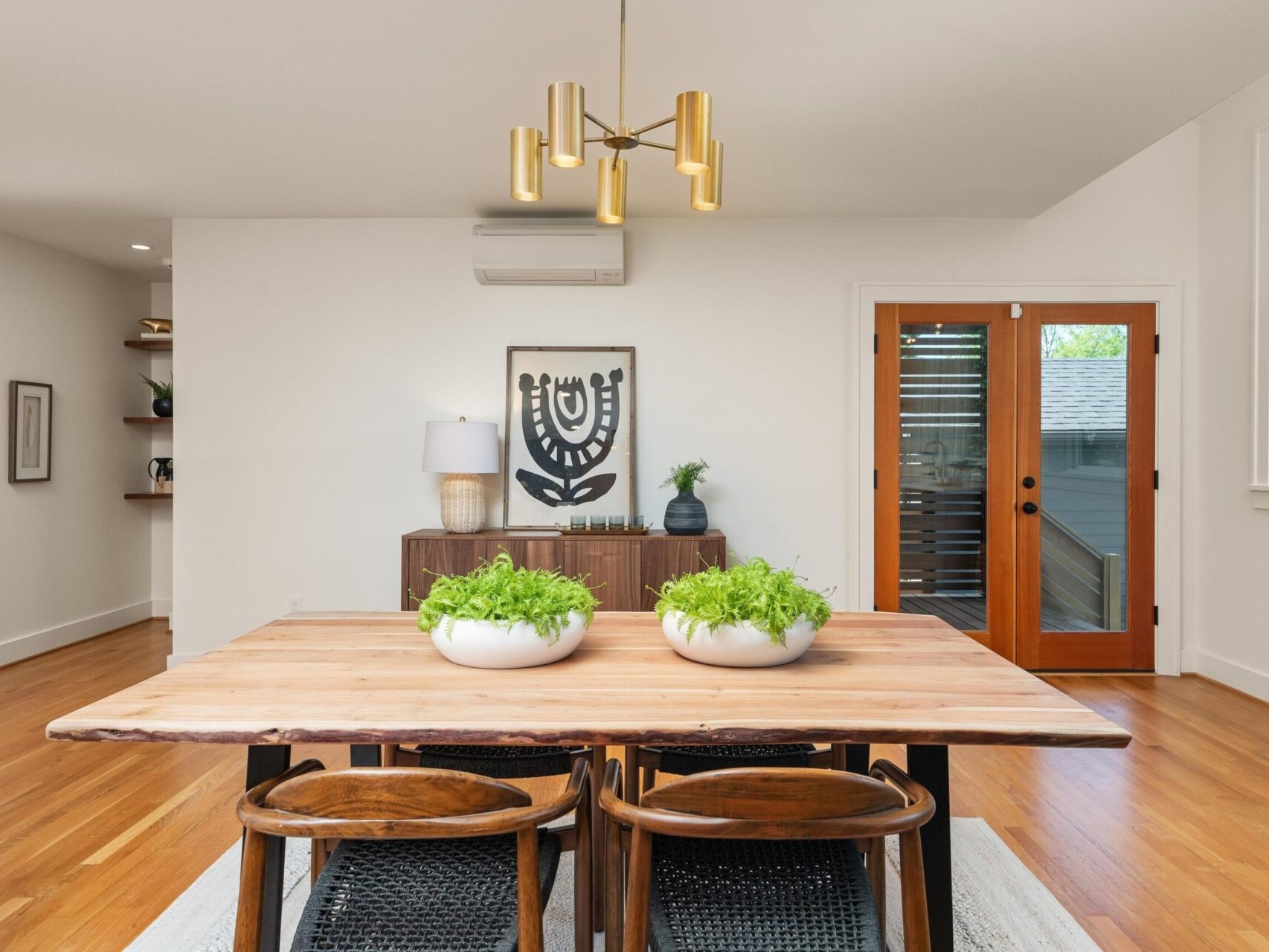 A modern dining room with a wooden table and four chairs. The table features two potted plants. A gold chandelier hangs above. In the background, theres a wooden cabinet, a framed art piece, and a glass door leading outside. Hardwood flooring is visible.