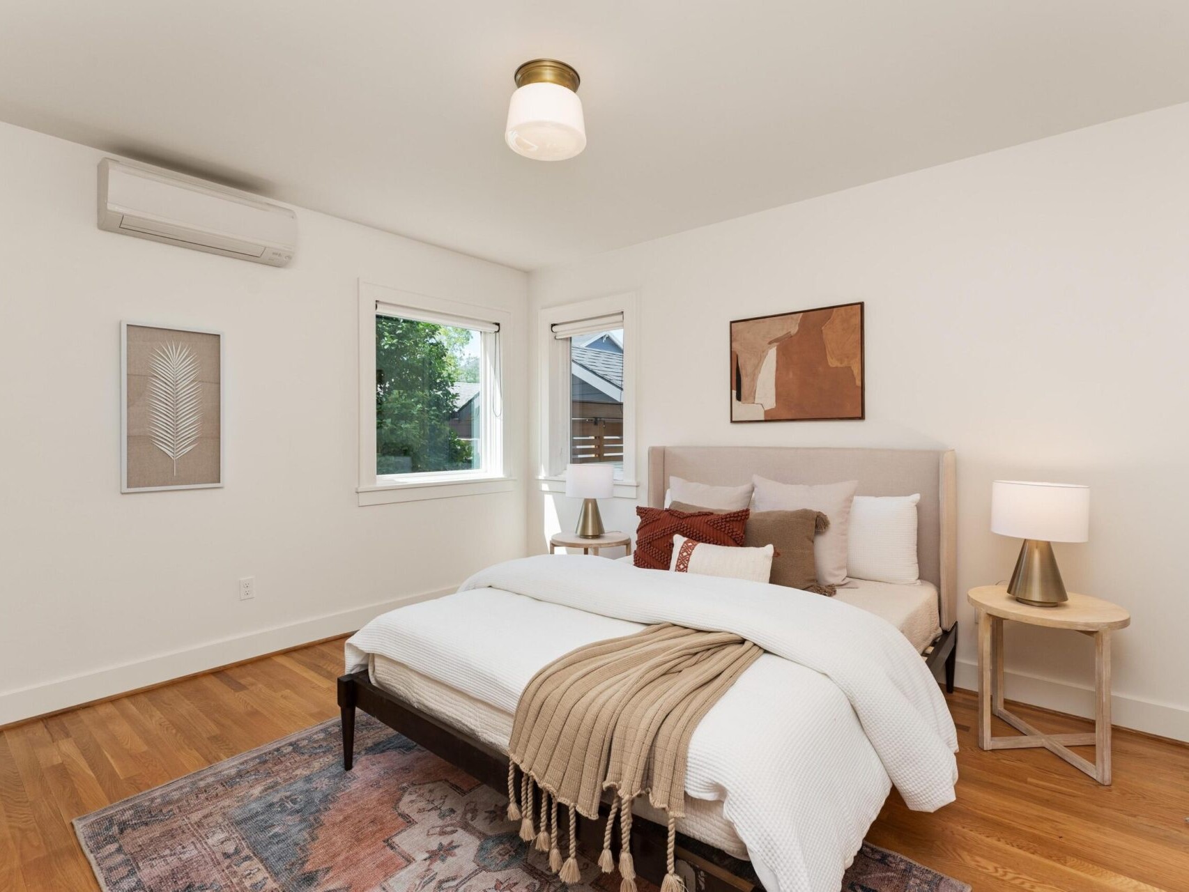 A cozy bedroom with a neatly made bed featuring white bedding and decorative pillows. A small side table with a lamp is beside the bed. Art hangs on the light-colored walls, and a window offers a view outside. Hardwood floors add warmth.